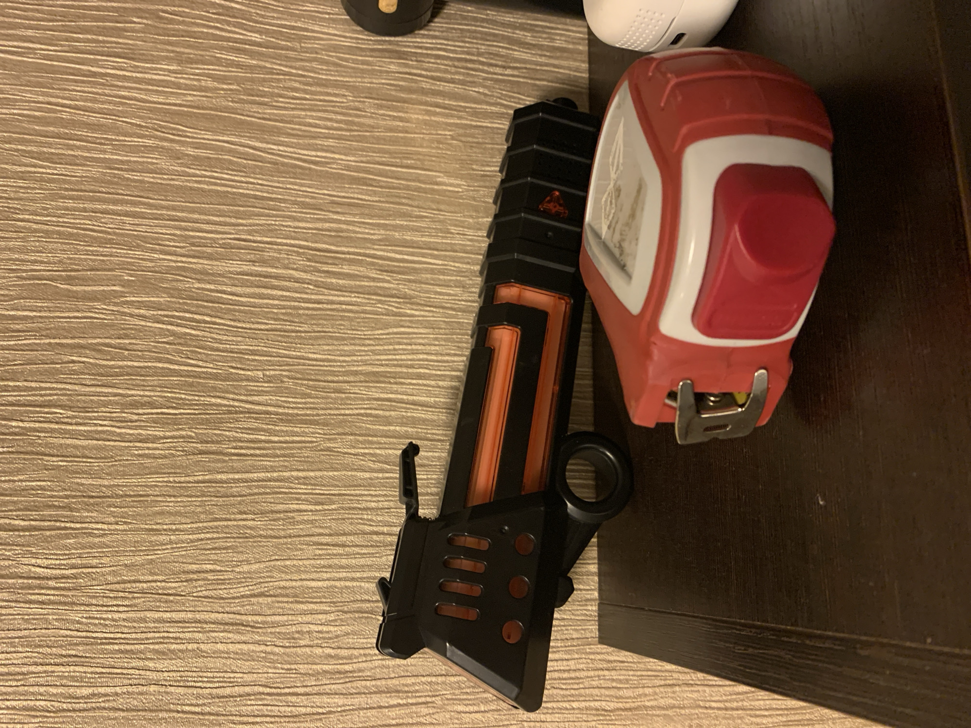 A toy gun and a tape measure sit on a dark wooden surface in front of a beige, textured wall. The toy gun is black with orange accents and has a futuristic design. The tape measure is red and white and is positioned in the foreground. The scene is indoors, with no people or animals present. The lighting is dim, suggesting it may be evening or a poorly lit room.