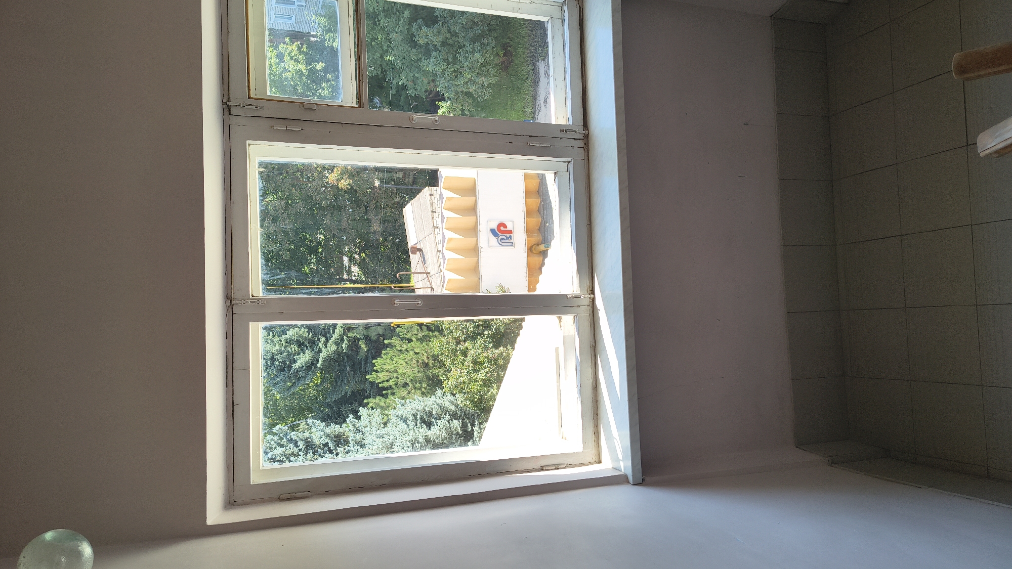 A wide shot, taken from a slightly low angle, shows a view through a multi-paned window. The window frame is painted white and appears somewhat aged. The left pane looks out onto a dense growth of evergreen trees, with a bright white structure visible below them, possibly a building or roof. The middle and right panes offer a view of a building with a white facade decorated with an emblem featuring red and blue circular shapes, and what appears to be a stylized "RP" or similar lettering. The building has a corrugated roof. More trees and foliage are visible behind and to the sides of this building. The interior wall on the left is painted a light pinkish-grey. The window sill is light grey with a marble-like pattern. Below the window sill, the wall is tiled with square, light grey tiles. The floor is also tiled with the same grey tiles. A portion of what appears to be wooden furniture is visible in the lower right corner of the frame. The overall lighting suggests a bright, daytime scene, with sunlight casting distinct shadows on the window sill. The scene is indoors, looking outwards. The location is identified as Mikhaylovka, Russia.