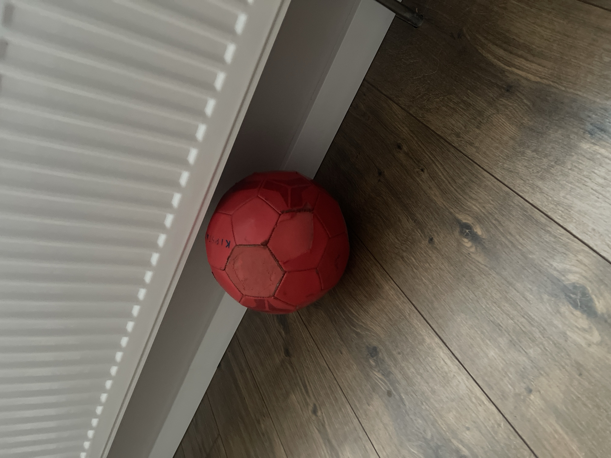 A red, multi-panel ball rests on a dark wood floor, positioned near the base of a white radiator. The ball has the brand name "Kipsta" partially visible in white lettering. The radiator features vertical fins and casts a shadow on the wall behind it, creating a pattern of repeating dashes of light. The scene is indoors, likely in a domestic setting, with no people or animals present. The lighting suggests an indoor environment with artificial or indirect natural light. The location in Molokovo, Russia, is noted, but there are no visual elements in the image to confirm this specific location.