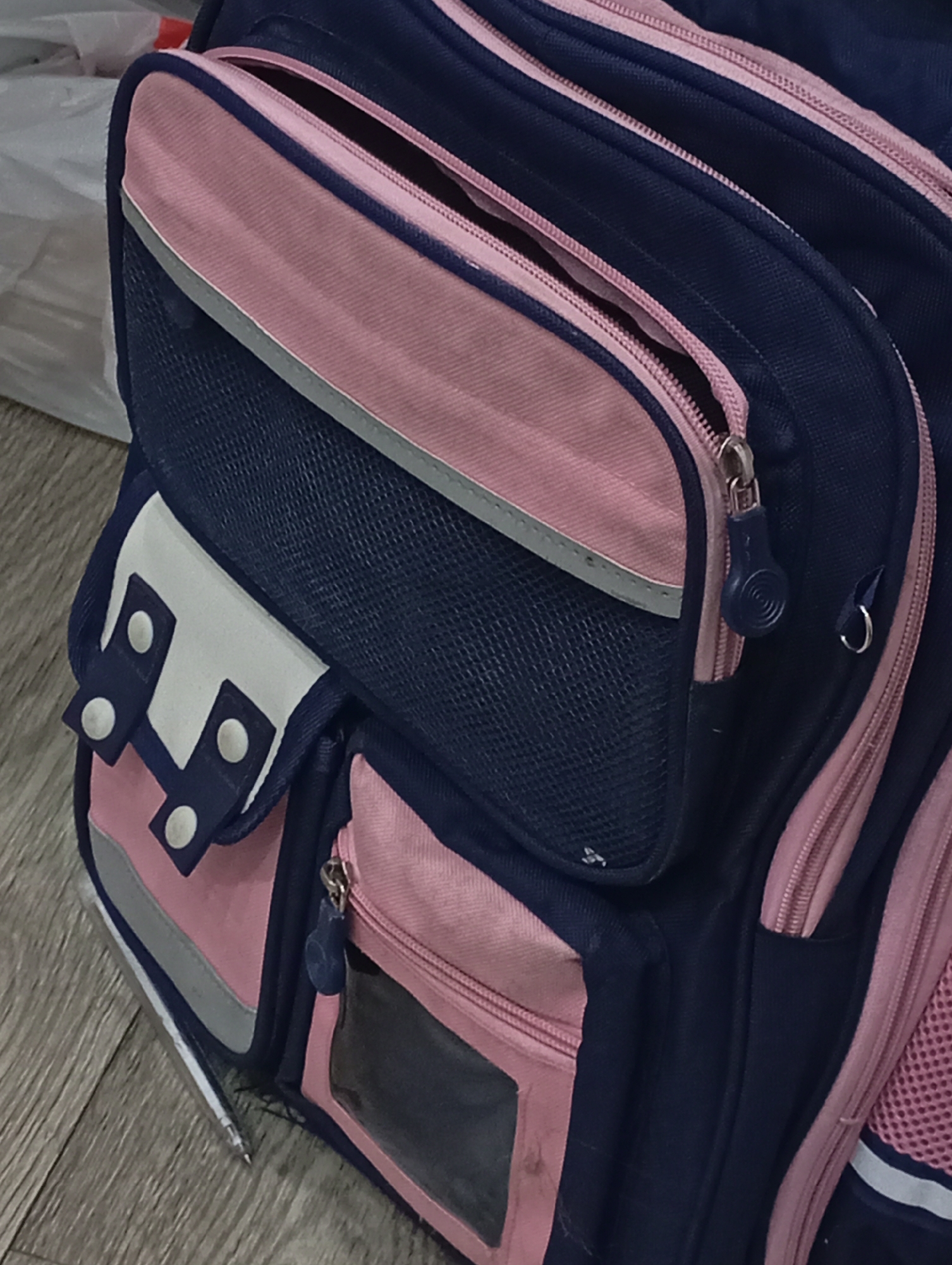 The image shows a blue and pink backpack with multiple pockets and zippers. The bag is resting on a wooden floor. A white plastic bag is visible in the upper left corner. A pen is visible in the left side pocket. The location is not discernible from the image.