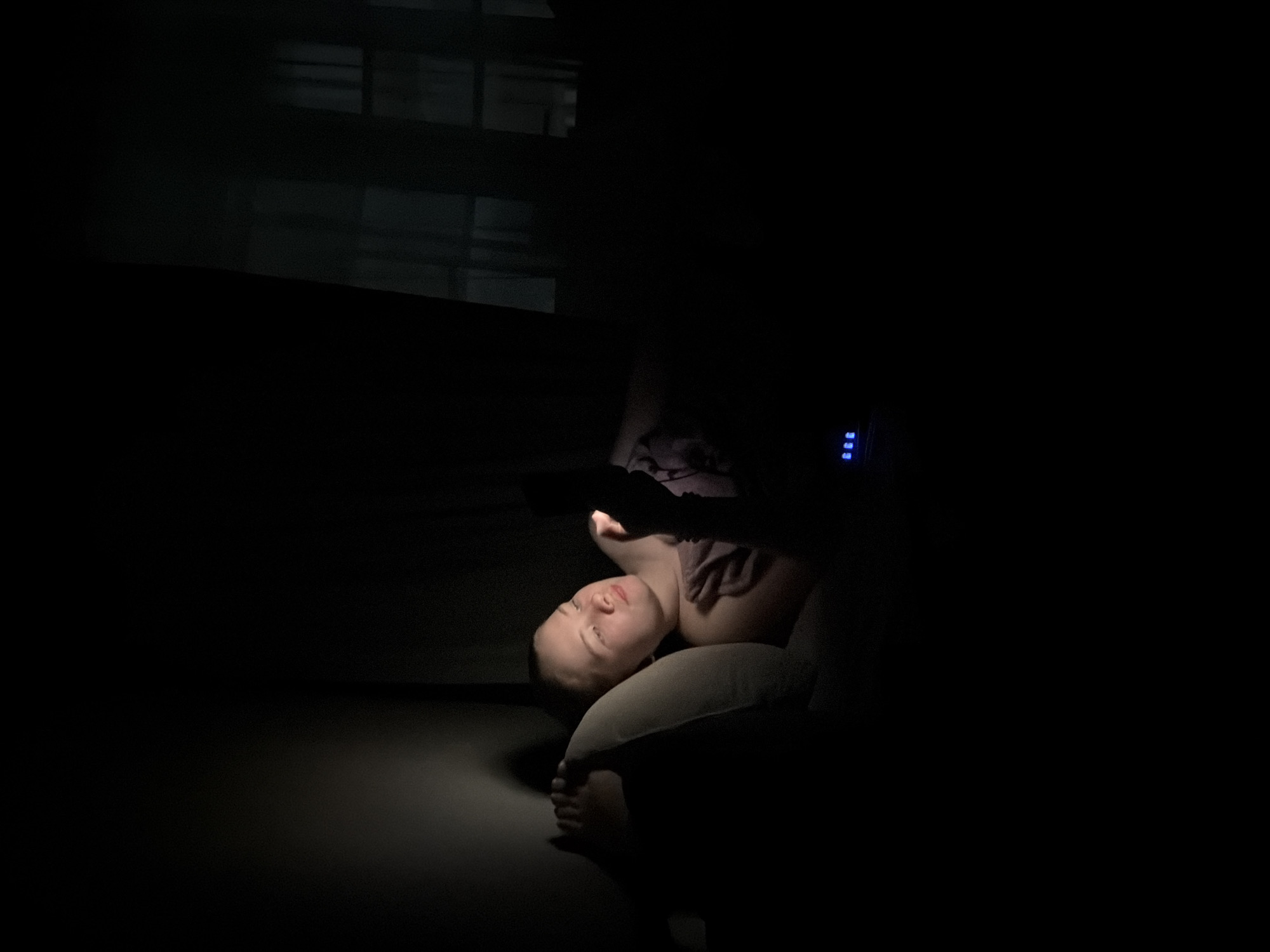 The image shows a person lying down in a dark room, illuminated primarily by the screen of a handheld electronic device, likely a smartphone. The person's head rests on a light-colored pillow or cushion, and their face, neck, shoulder, and one bare foot are visible. They are looking directly at the glowing device, which they hold in their hands, suggesting engagement with its content. Their expression is neutral, appearing focused or relaxed.
The setting is an indoor space, strongly implying nighttime due to the deep darkness that envelops most of the room. The device's light creates dramatic contrasts, brightly illuminating the subject while casting the surroundings into deep shadow. In the upper right portion of the image, a faint blue digital display with multiple segments (indistinct to read specific numbers) emits a soft glow from an unseen appliance. No other significant objects or clear textual elements are discernible in the dimly lit environment. The overall atmosphere conveys a sense of quiet, personal activity, consistent with using an electronic device in a private space at night. The location context provided is Chaikovsky, Russia.