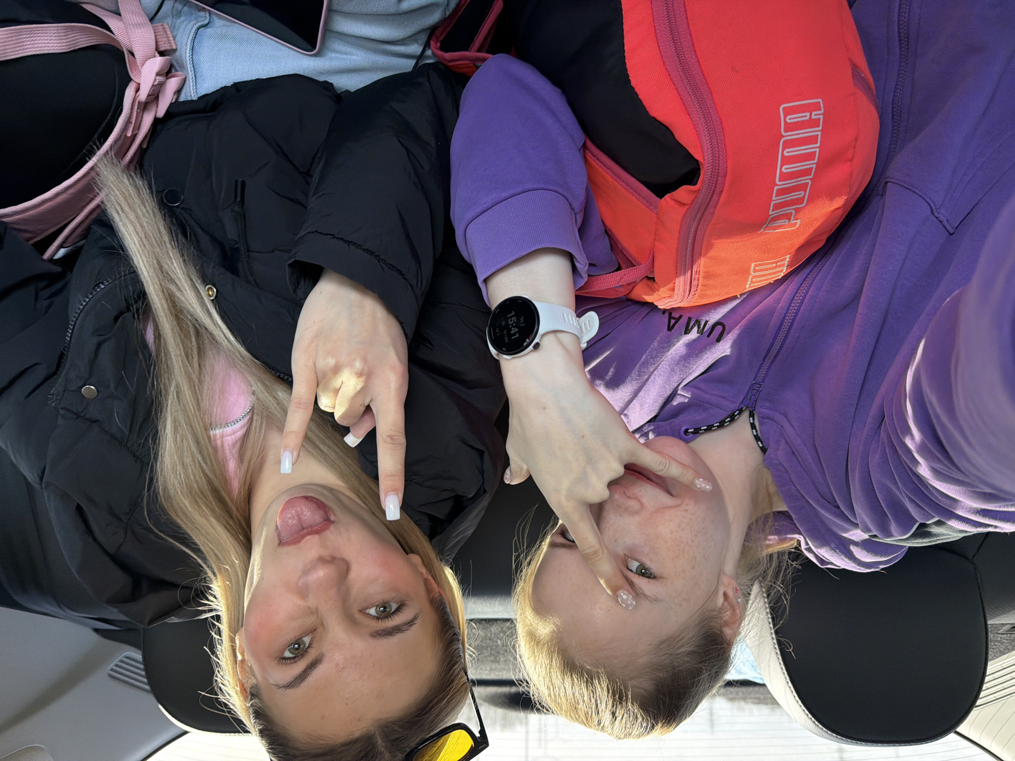 Two young women pose for a selfie in what appears to be a car. The woman on the left has blonde hair and is wearing a black jacket over a pink top. She is sticking out her tongue and making a hand gesture. The woman on the right has blonde hair and is wearing a purple hoodie. She is also making a hand gesture and wearing a white smartwatch. A pink and black bag is visible behind the woman on the left, and a red bag with "Puma" printed on it is behind the woman on the right. The location is unknown and cannot be determined from the image.
