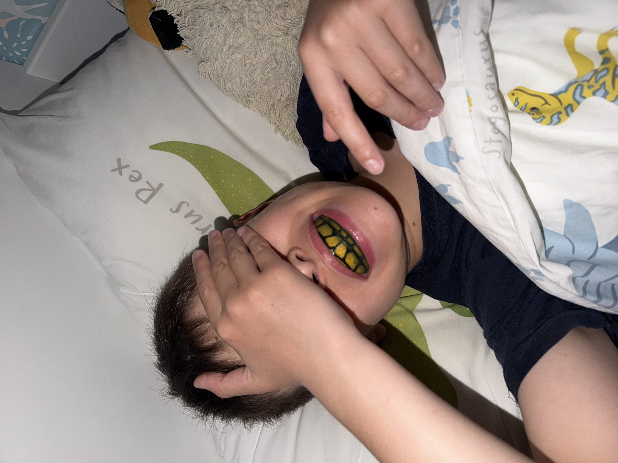 A child lies in bed with a blanket pulled up, partially obscuring their face. The child is wearing a dark blue or black shirt. A hand is covering part of the face. The child is wearing a novelty mouthpiece with a black and yellow snake-like pattern. The bed has a white pillowcase with a green dinosaur tail and the words "rus Rex". The blanket also has dinosaur patterns. The location is presumably indoors in a bedroom. There is no information to ascertain whether the location is in the city of Votkinsk, Russia.