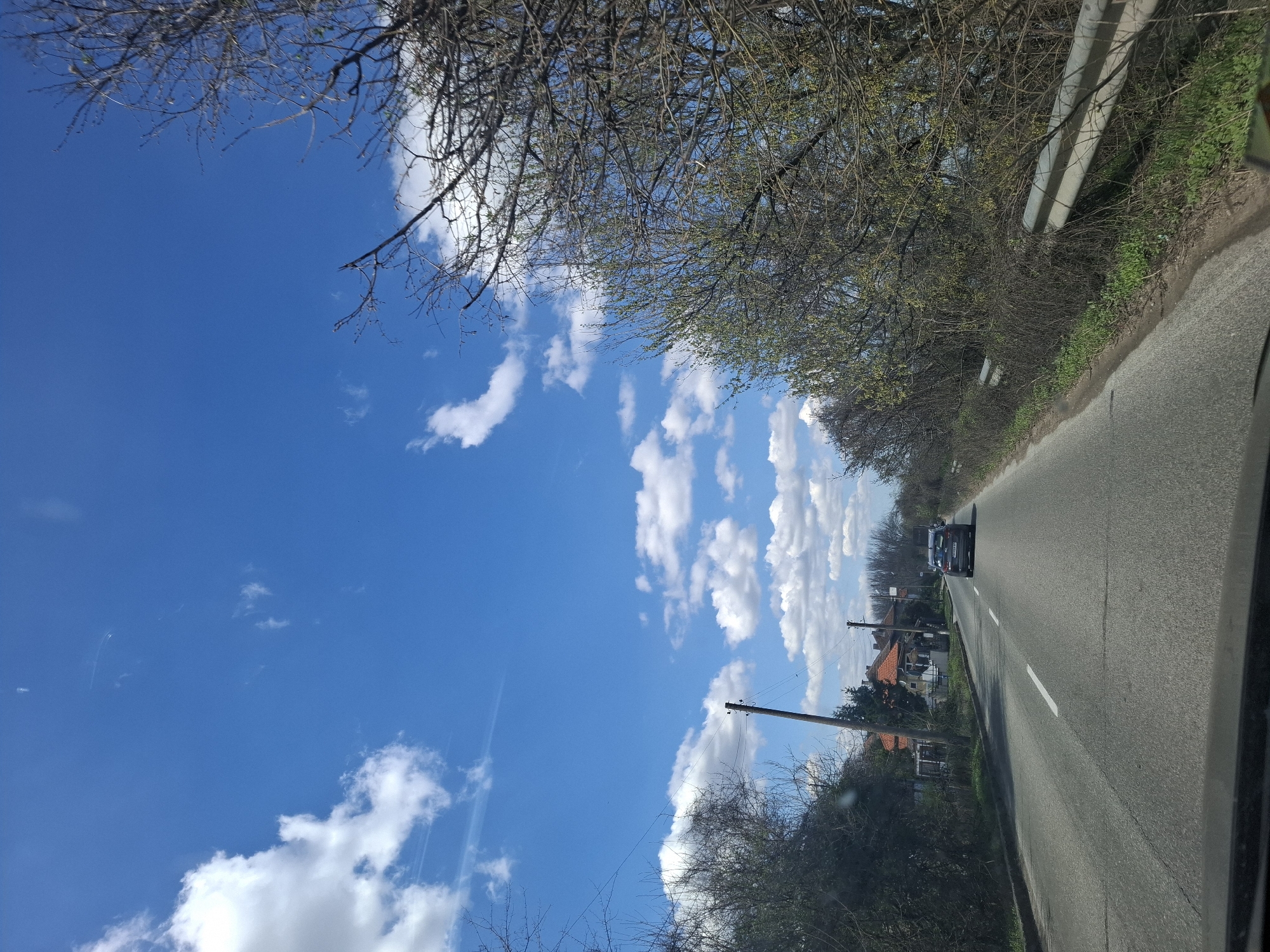 The image depicts a road in Botevo, Bulgaria, as viewed from inside a moving vehicle. The sky is blue with scattered white clouds. Bare tree branches are visible at the top left of the image. A road curves slightly to the right, with a guardrail along the right side. A dark car is traveling in the same direction ahead on the road. Houses with orange tile roofs and power lines are visible to the left of the road. The road has a single solid white line marking the center. The dashboard of the vehicle taking the picture is visible at the bottom right.