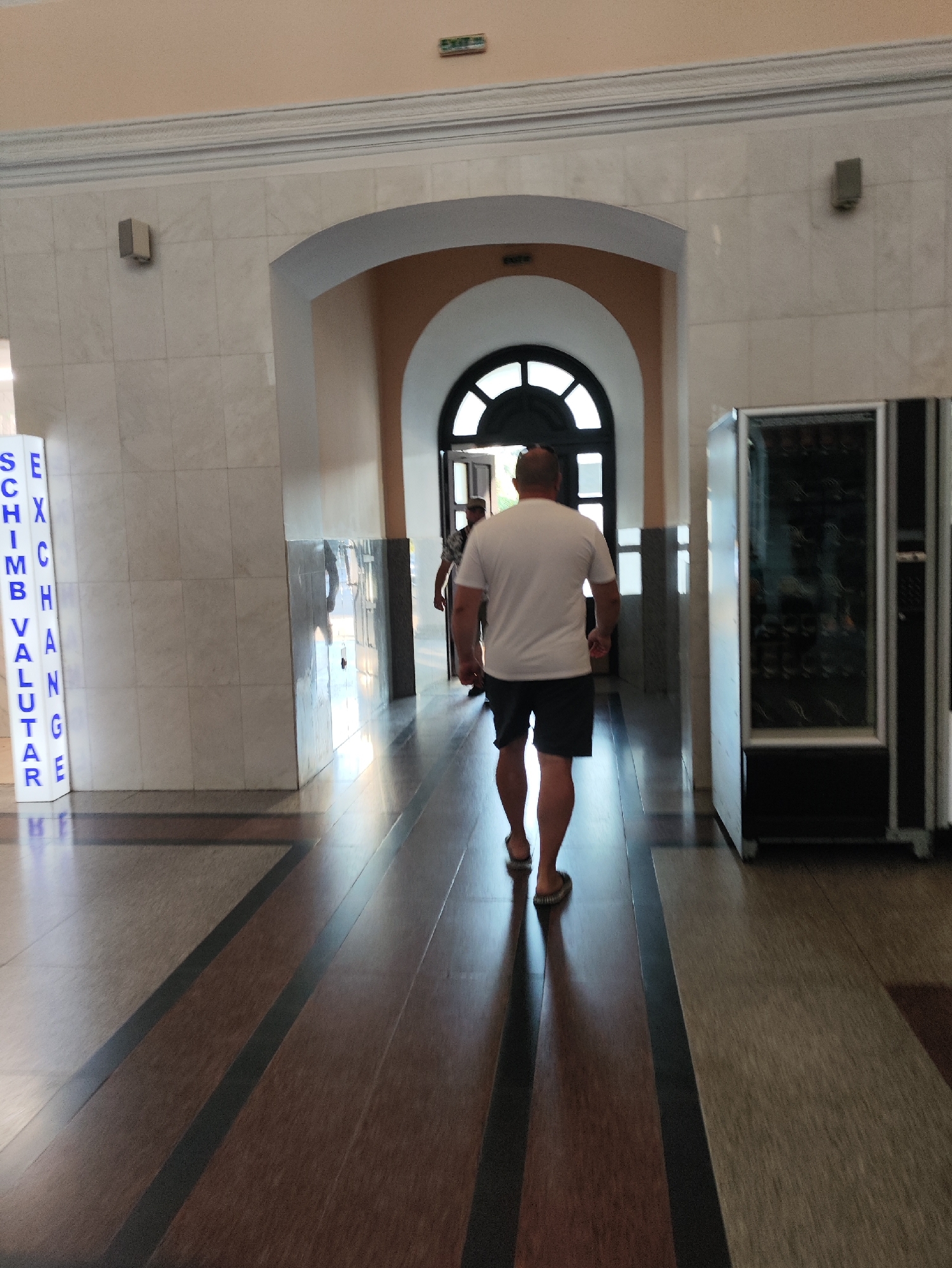 A bald man in a white t-shirt and dark shorts walks away from the camera down a polished hallway. The hallway has dark wood floors with lighter wood and black inlaid geometric patterns. To the left, a vertical sign reads "SCHIMB VALUTAR EXCHANGE" in blue letters on a white background. To the right, a large, tall, silver and black vending machine is visible. The man is walking towards an arched doorway with a black, arched window. The walls are lined with light-colored tiles, and decorative molding runs along the top of the walls. A green "EXIT" sign is visible on the ceiling. The scene appears to be an interior hallway, possibly in a public building such as an airport or train station, and could be located in Chișinău, Moldova, given the Romanian text on the sign.