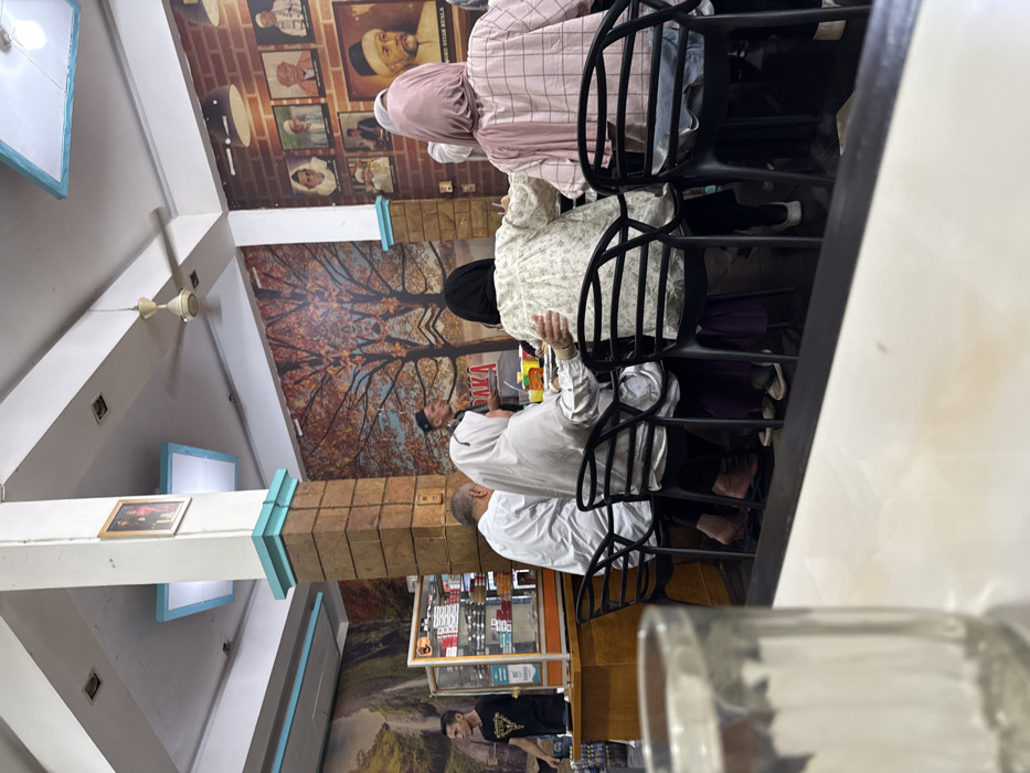 An interior view depicts an indoor dining establishment in the city of Payameuneng, Indonesia. In the immediate foreground, a blurred clear glass tumbler rests on a light-colored surface.
Five individuals are seated at a long counter or table in the midground, with their backs partially or fully to the viewer, occupying black, slatted chairs. From left to right: one individual wears a white shirt and dark sandals; another wears a white headscarf and white top with dark sandals, their arm extended towards the person next to them; a third wears a light-colored, floral-patterned long-sleeved top and dark pants with light shoes; and a fourth wears a pink plaid long-sleeved top and a light pink headscarf over jeans. The fifth individual is mostly obscured.
To the left, behind the seated group, a male staff member in a black t-shirt with a gold graphic stands behind a wooden counter featuring a glass display case filled with various packaged goods. In the central background, another male staff member wearing a dark cap and shirt stands facing the seated individuals.
The environment includes a white ceiling with recessed light panels framed in light blue, and a beige ceiling fan. Two prominent square pillars, clad in a brown brick-like texture with light blue trim at the top and base, extend from floor to ceiling; one displays a small framed picture. The walls feature distinct murals: the left wall depicts a waterfall and green landscape; the central back wall displays a mirrored image of autumnal trees with reddish-brown foliage. The right wall is covered in a reddish-brown brick-patterned wallpaper adorned with multiple framed portraits of men, some in traditional attire, including a large portrait of a bearded man with "SYIAH KUALA" text visible below it.