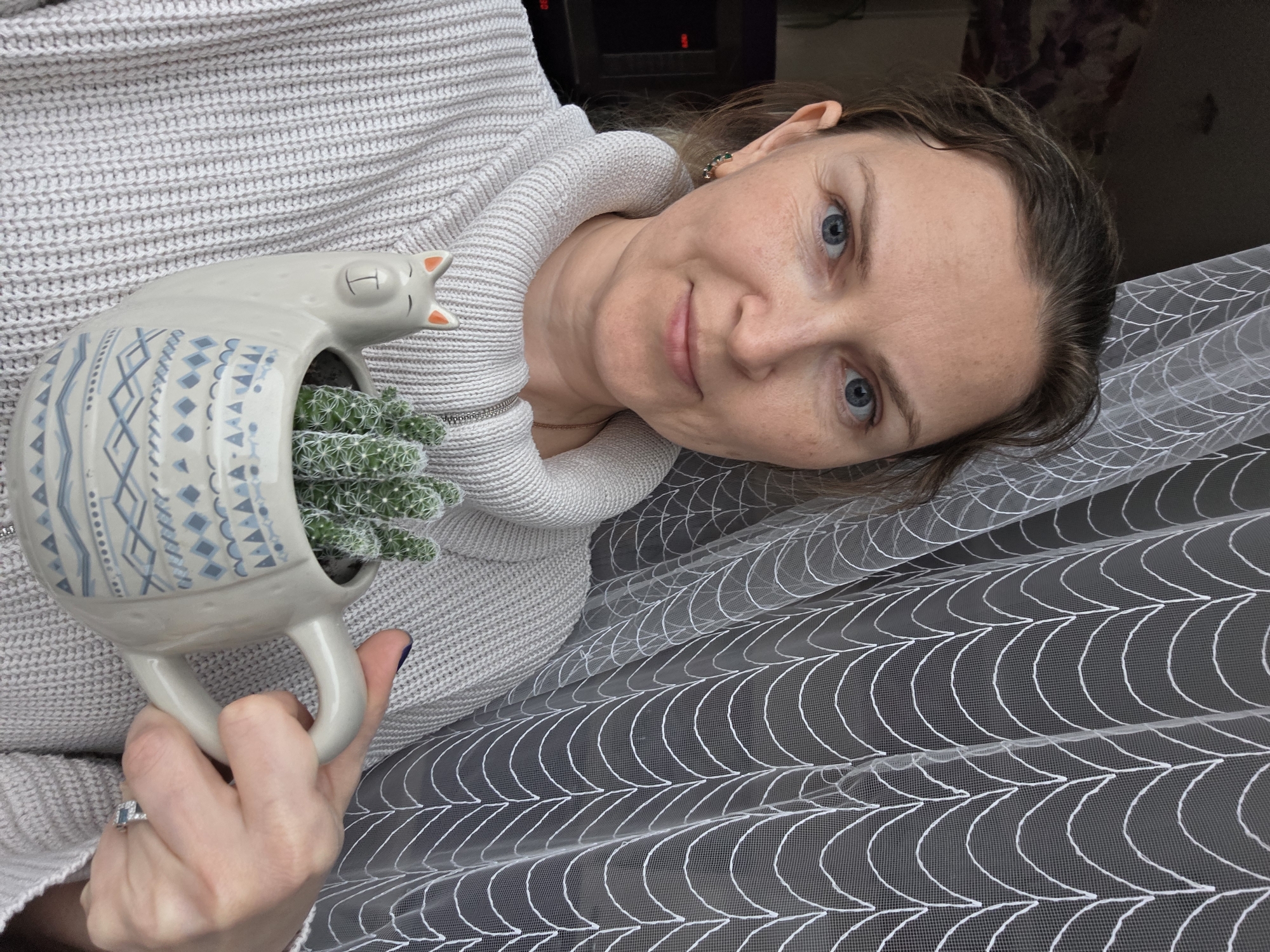 A woman, likely in Chaikovsky, Russia, is featured in a close-up, upward-angled portrait shot, looking directly at the camera with a gentle, slight smile. She has light skin, blue eyes, and light brown hair, and is wearing a light grey knitted sweater with a ribbed texture.
She holds a decorative ceramic planter in her right hand. The planter is light-colored, possibly beige or off-white, and shaped like a sleeping cat or llama with small pointed ears and closed eyes. It is adorned with blue and white geometric patterns. Inside the planter are several small, green cacti or succulents. A ring with a light blue gemstone is visible on her left ring finger, and she wears small, possibly green, earrings in her right ear.
The background consists of a sheer white curtain or net fabric, featuring a distinctive pattern of overlapping white semicircles or arches, suggesting an indoor setting near a window. A darker, indistinct background element is visible behind her head. The overall lighting suggests it is daytime. There is no visible text in the image.