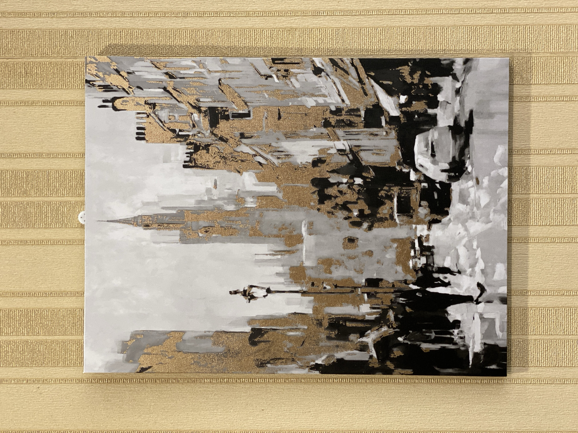 This is a photograph of a square canvas painting with an abstract design. The painting features a monochromatic palette of black, white, and various shades of gray, accented with gold leaf. The style is impressionistic and somewhat abstract, with brushstrokes visible that create a textured and dynamic effect.
The subject matter appears to be an urban landscape, possibly a cityscape, with implied architectural elements such as buildings and possibly a tower or spire in the center-left. The gold leaf is applied in irregular patches and streaks, catching the light and adding a metallic sheen that contrasts with the muted tones of the rest of the painting. The composition is somewhat fragmented and layered, suggesting depth and movement within the abstract forms.
The painting is hung on a textured wall, which has a subtle, horizontal ribbed pattern and a light tan or beige color. The wall texture provides a tactile background that complements the visual texture of the artwork.
There are no visible people or distinct objects, and the abstract nature of the painting makes it difficult to pinpoint a specific activity or event. The lighting in the photograph appears to be general indoor lighting, casting a soft shadow on the wall behind the top edge of the canvas. No specific time of day or weather conditions are suggested by the artwork itself. There is no visible text within the painting or on the surrounding wall. The overall impression is one of modern, sophisticated abstract art.