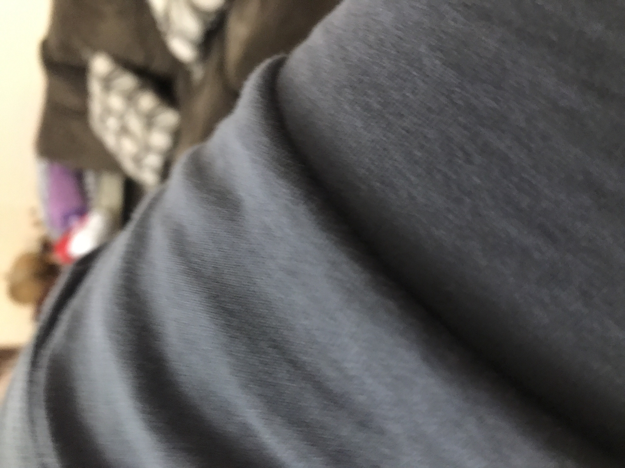 The image primarily focuses on a grey fabric, possibly clothing, filling the majority of the frame. The texture is visible, showing subtle creases and variations in the fabric's surface. In the blurred background, there are hints of a brown object with a patterned element (possibly a cushion or pillow) and glimpses of other indistinct items, suggesting a domestic interior. The image provides no indication of being in Yerevan, Armenia, or any other specific location.