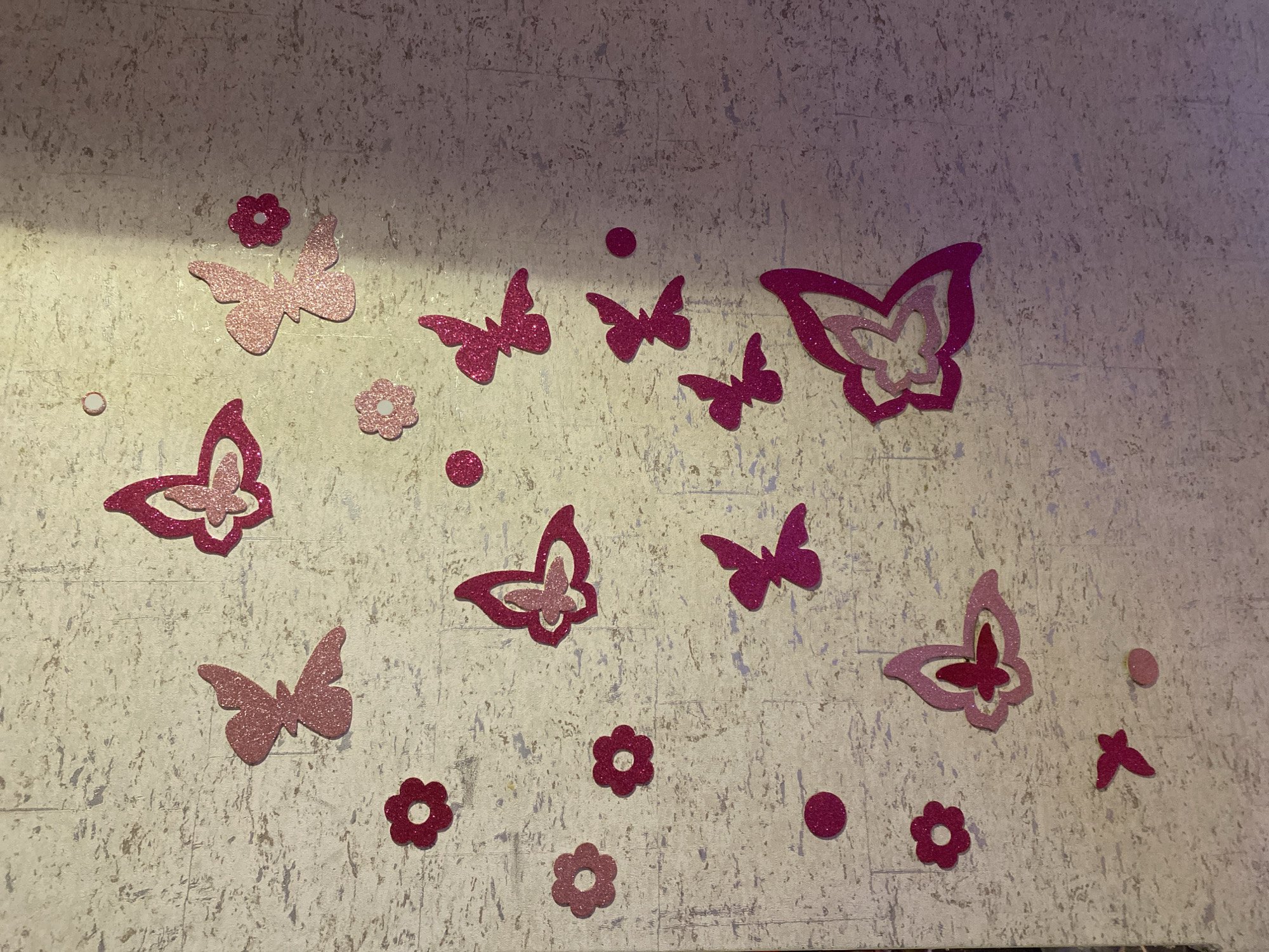 The image shows a wall decorated with pink glittery butterfly and flower stickers. The butterflies vary in size and shade of pink, with some having intricate patterns and others being solid colors. The flower stickers are also pink and have a simple, stylized design. The wall itself has a speckled texture, appearing to be made of a material like corkboard or textured wallpaper. The lighting in the image creates shadows, suggesting an indoor setting. There is no visible text in the image. The overall impression is of a whimsical and decorative wall, likely in a child's room or a creative space.