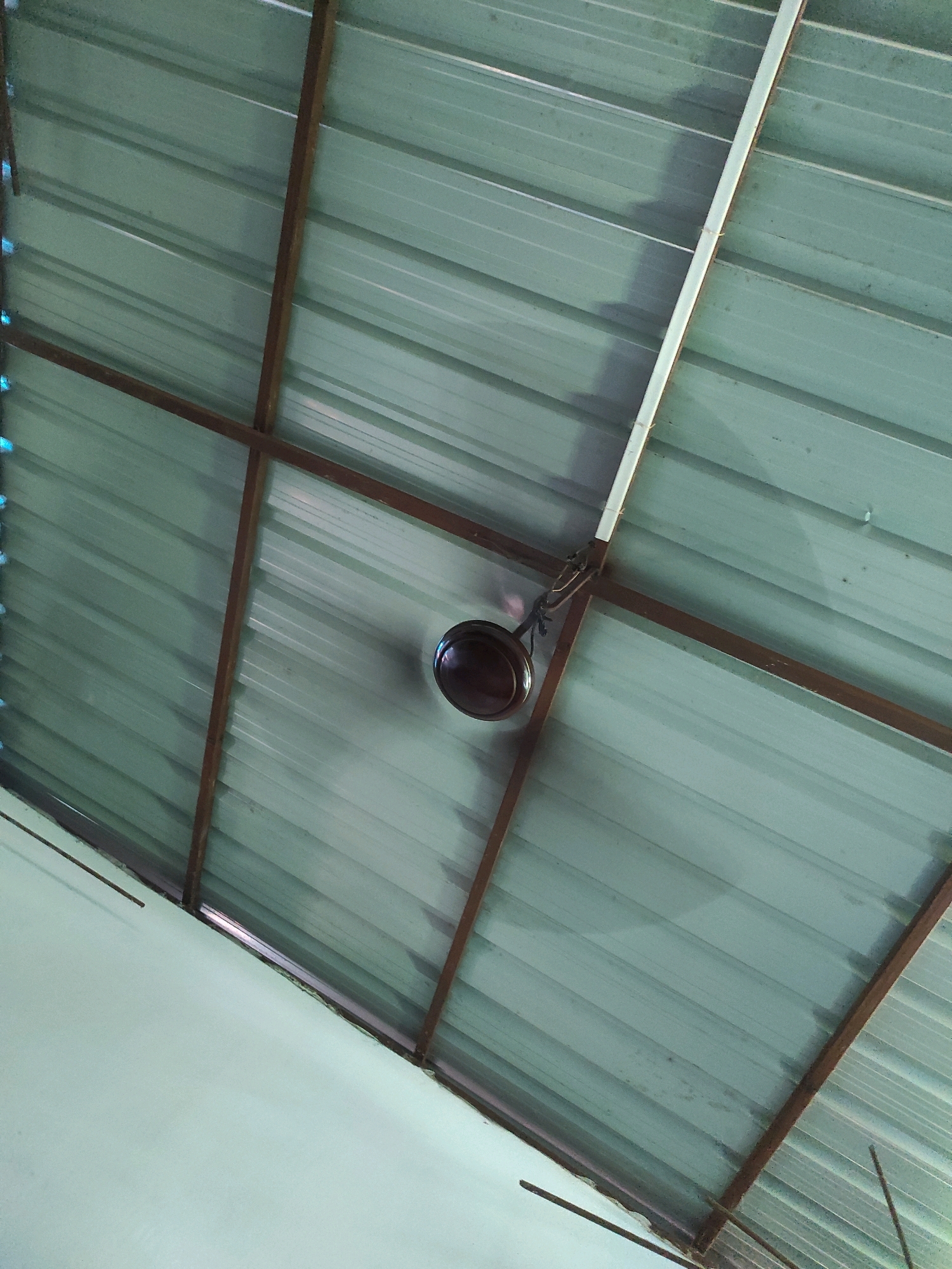 A low-angle shot looks up at a metallic, corrugated roof supported by a metal frame. A ceiling fan with a dark brown motor is attached to the roof. The overall color of the roof is a muted green. A wall, painted a light green, is visible at the bottom left of the image. The scene appears to be indoors, possibly in a residential or commercial building in Islampur, India. There are no people or animals visible in the frame. The lighting suggests daytime, with some shadows cast on the roof.