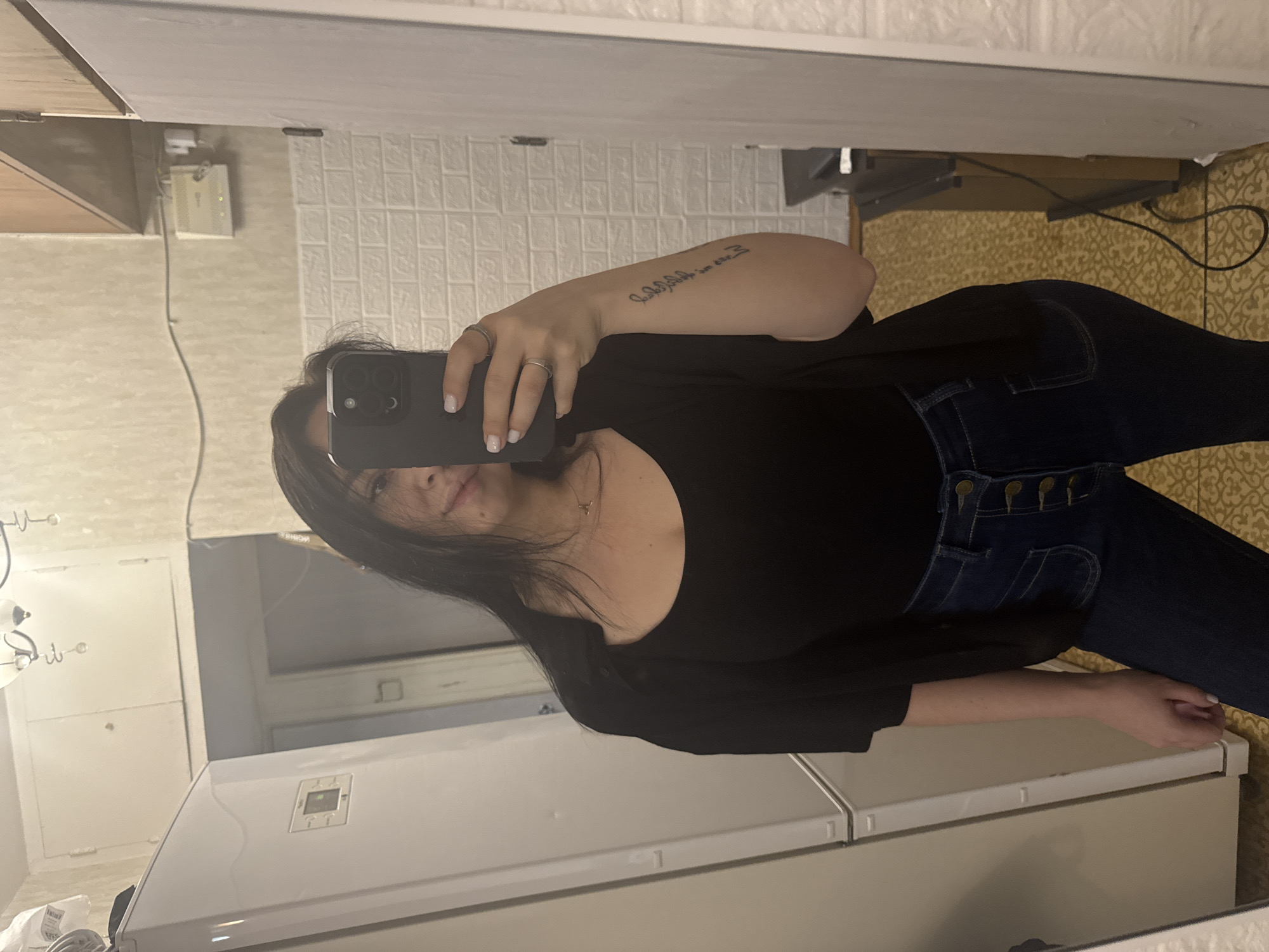A woman stands in a room, visible in a mirror's reflection. She is taking a selfie with a smartphone. She has long, dark hair and is wearing a black top and dark denim jeans with button accents. A tattoo is visible on her left arm. The room includes a white tiled wall, a brown cabinet, and a patterned floor. Reflecting in the mirror is a white refrigerator. The image was taken in a domestic setting, consistent with an apartment or home environment. The room appears to be a kitchen or dining area. It is not possible to confirm the location in Mytishchi, Russia based only on the provided image.