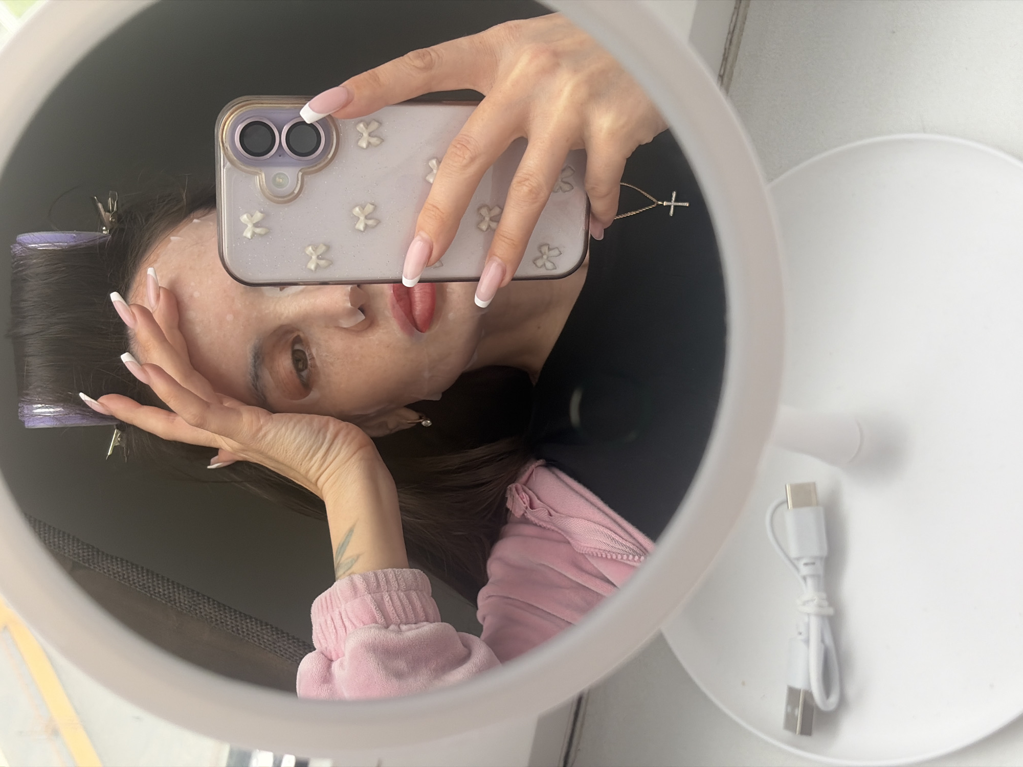 A reflection in a ring light mirror shows a person taking a selfie with a smartphone. The person has a face mask on, hair clips in their hair, and is wearing a pink sweatshirt. They have long nails with white tips. The smartphone has a decorative case with small white flowers and a small gold cross charm hanging from it. A white USB charging cable rests on the base of the ring light. The location is Unknown, Russia.