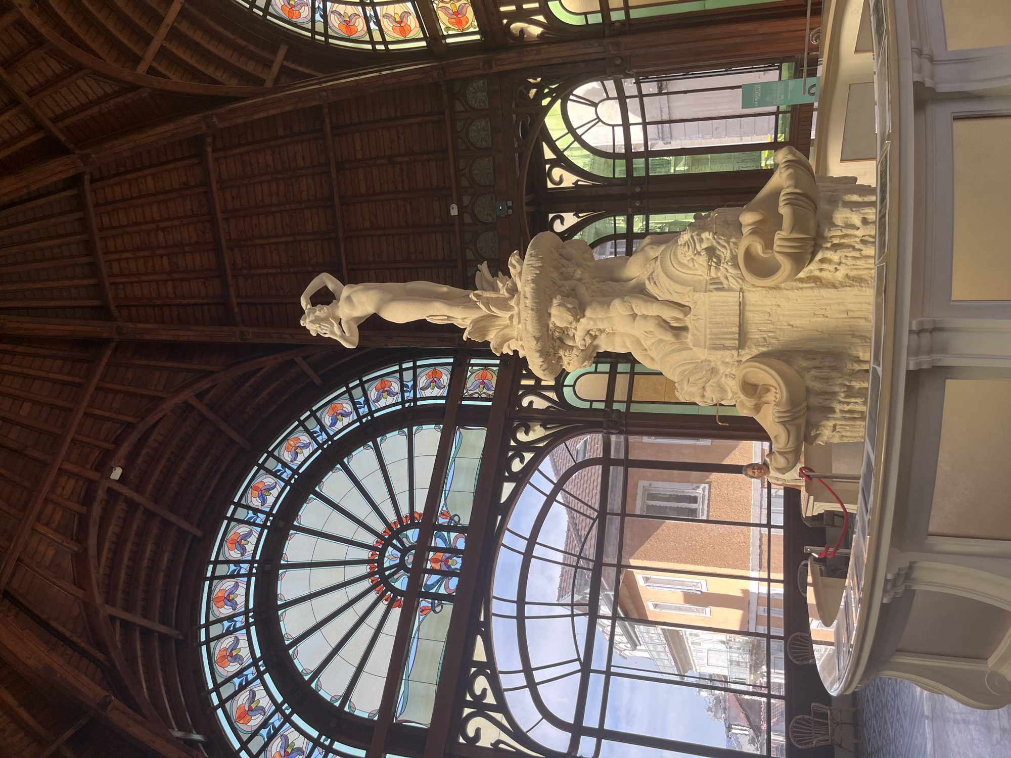 The image depicts the interior of the Palais Lumière in Évian-les-Bains, France, a former thermal spa now serving as a cultural center and exhibition space. Dominating the foreground is a large, ornate, off-white stone fountain sculpture. The fountain features a central figure of a nude woman with her arms raised, standing atop a tiered base adorned with cherubs and architectural motifs, including what appear to be carved shells or urns.
Behind the fountain, a series of tall, arched windows with stained-glass accents allow natural light to filter into the space. The stained glass features intricate patterns of floral designs in shades of red, orange, purple, and blue. The windows look out onto a street scene with buildings, including a prominent orange building with balconies and windows, under a bright sky.
The interior structure is characterized by exposed wooden beams forming a vaulted ceiling, characteristic of Belle Époque architecture. Wooden decorative elements and frames are visible throughout the space, surrounding the windows and supporting the ceiling.
In the middle ground, a curved, light-colored counter or railing encloses the fountain area. A red rope barrier is visible along this counter. A woman with short dark hair, wearing a light-colored top, stands behind the counter, partially obscured by the fountain sculpture. In the background, through the windows, several empty wicker chairs are visible, suggesting seating areas.
The overall impression is one of grandeur and historical significance, showcasing the architectural beauty of the Palais Lumière and its centerpiece fountain. The lighting is bright, with natural light coming from the windows, highlighting the details of the sculpture and the stained glass.