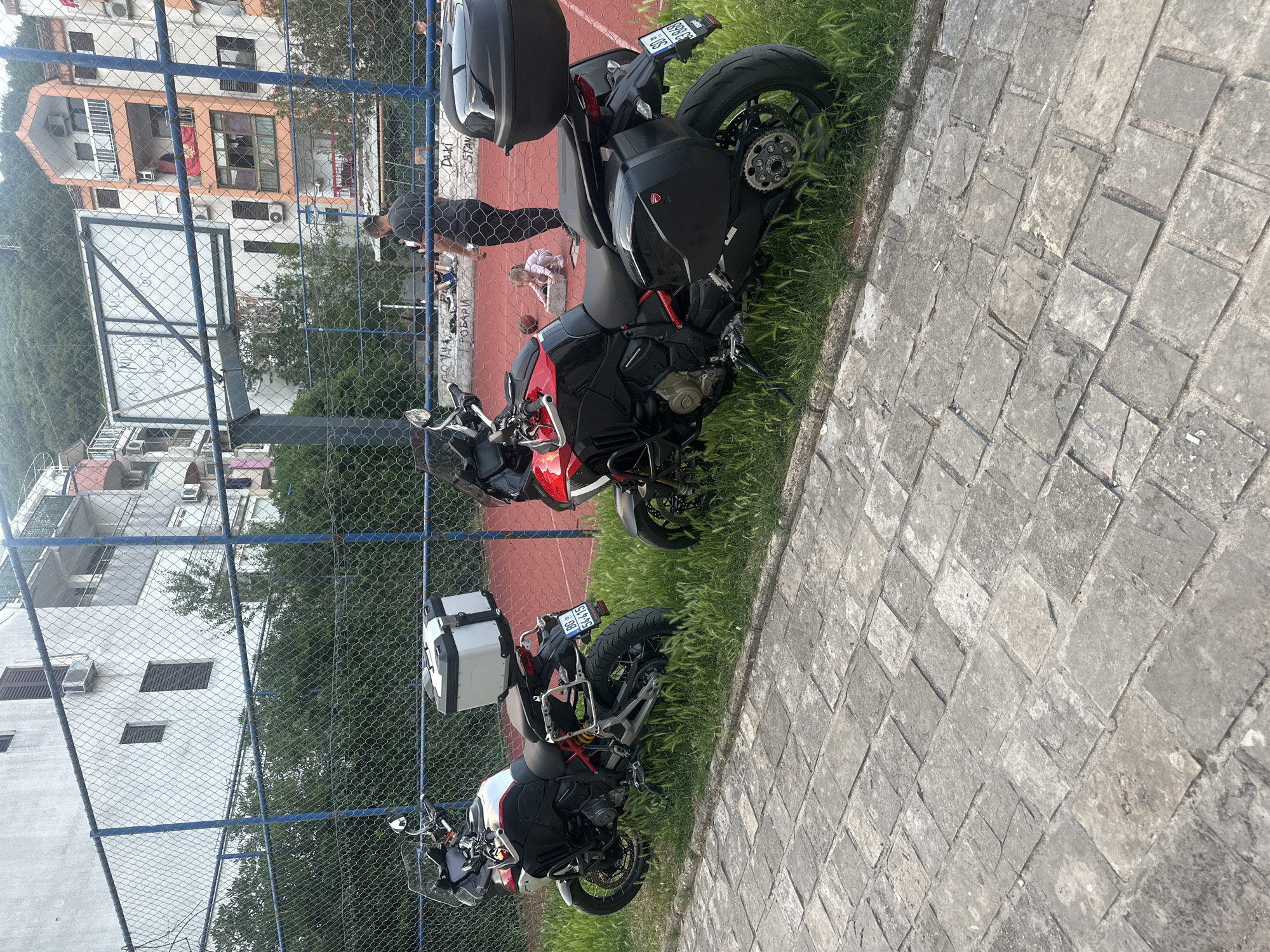 An outdoor, side-angle shot captures two touring motorcycles parked on a grassy verge next to a paved area, with a sports court and buildings in the background, all viewed through a chain-link fence.
In the immediate foreground, two motorcycles are prominent. The closest is a red and black adventure-style motorcycle, equipped with black hard panniers and a matching top case. Its license plate, "SD 888 CS," is partially visible. Further back, a white and red adventure-style motorcycle is parked, also fitted with white hard panniers and a top case. Its license plate, "BG 544-Y5," shows "SRB" (Serbia) as the country code. Both bikes are parked on a narrow strip of green grass, adjacent to a grey stone-paved walkway.
Beyond the paved area and motorcycles, a blue-framed chain-link fence spans the scene. Behind this fence is a red-surfaced sports court, possibly for basketball or tennis. A basketball hoop with a white backboard is visible on the left. Several people are on the court: a man in a dark t-shirt is bending over, and a child is seated on the ground nearby. Text is partially visible on the wall behind the court, including "DAKI STAN," "PDBARIJ," and "TAXI."
In the background, multiple-story residential buildings with balconies and air conditioning units are seen. Green hills and trees rise behind the buildings, indicating a natural landscape. The lighting suggests it is daytime, likely morning or late afternoon, with clear weather. The presence of Serbian license plates, combined with the context of Budva, Montenegro, indicates these are visitors.