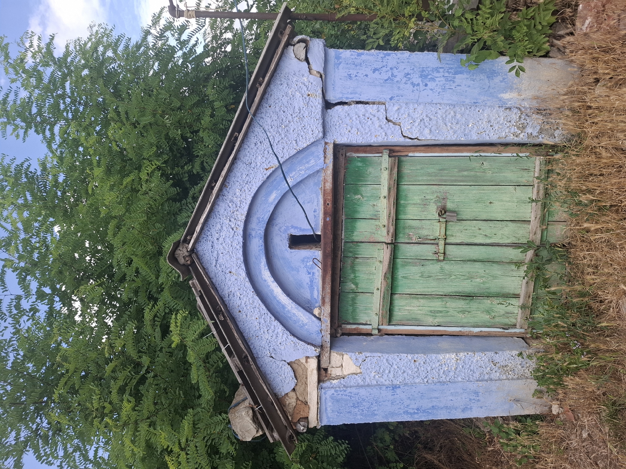 This is a vertical, eye-level medium shot of a dilapidated small structure with a blue, stucco exterior, and weathered green wooden double doors. The structure appears to be in a state of disrepair, with cracks in the stucco and exposed brickwork. The roofline is angled, with wooden beams extending outwards. A semi-circular archway is carved into the stucco above the doors, with a circular opening in its center.
The doors are made of horizontal planks of wood painted green and secured with a metal latch and a padlock. The wood is worn and faded, with some planks showing signs of rot. Green foliage from an overhanging tree covers the left side of the structure and the upper portion of the frame, and dry, golden grass grows on the right side. The sky, visible through the leaves, is blue with scattered white clouds, suggesting daytime with fair weather.
The overall impression is one of neglect and abandonment. The setting appears to be rural, possibly in a garden or a yard, given the surrounding vegetation. The image does not depict any people or animals, and there is no visible text. The location is identified as Chorna, Ukraine.