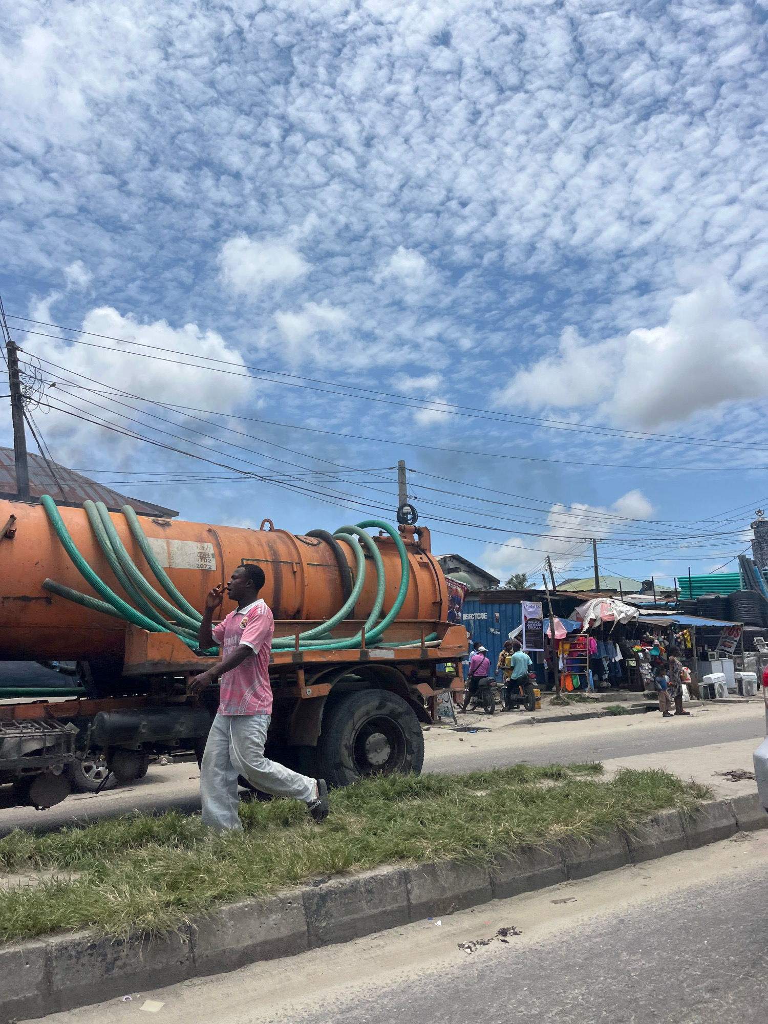 In Ado, Nigeria, a man in a pink t-shirt and grey pants walks on the side of a street beside an orange tanker truck. The tanker truck has several green hoses attached to it. In the background, there are market stalls with various goods displayed, including clothing and what appear to be appliances. People are seen browsing the stalls and riding motorcycles. The sky above is blue with scattered white clouds. Power lines are visible overhead, supported by utility poles. The scene depicts a busy street market environment.