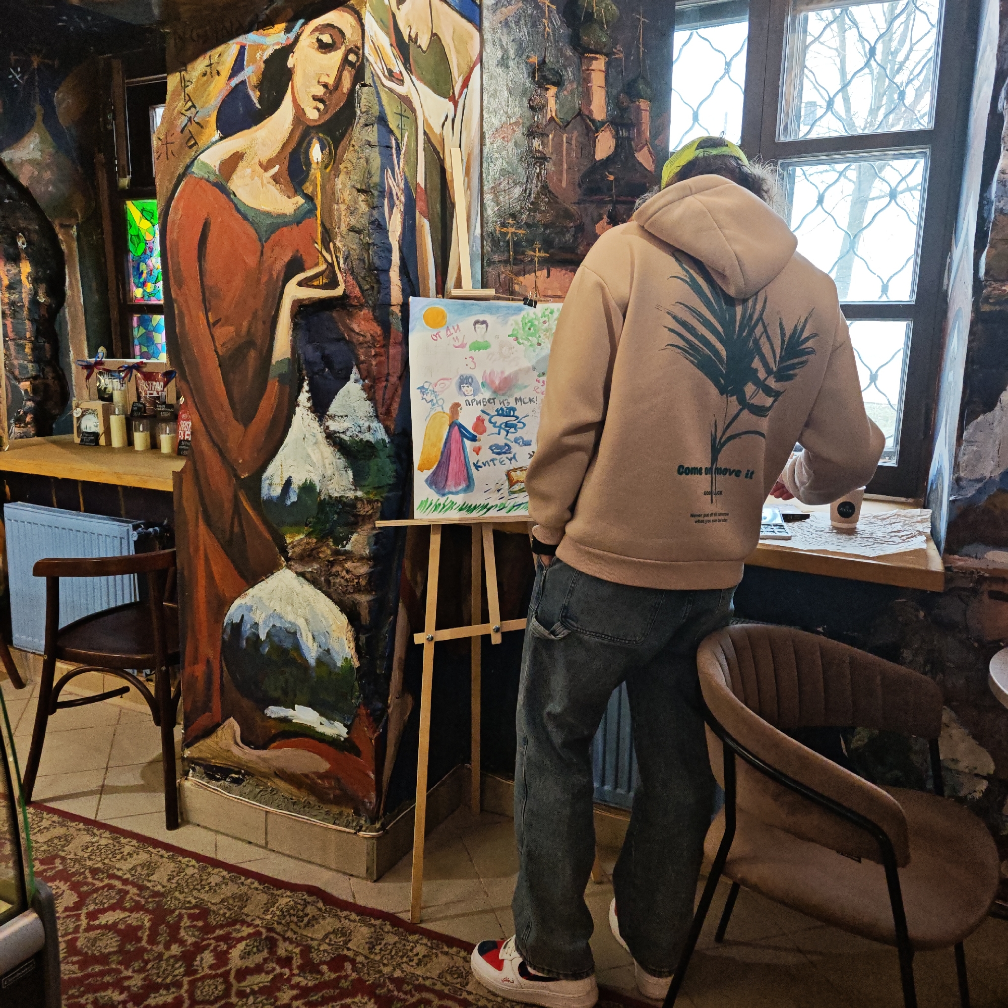 A person wearing a tan hoodie with a green palm tree design and the phrase "Come on move it" stands in front of a painting on an easel. The painting appears to be a child-like drawing, possibly in watercolor, with Russian text. To the left of the easel, a large painting of a woman holding a candle is on the wall. There is a window behind the person, and a cup sits on the windowsill. In the background, a bar area with bottles and other items is visible. Two chairs are positioned on either side of the easel. The location is Rostov, Russia.