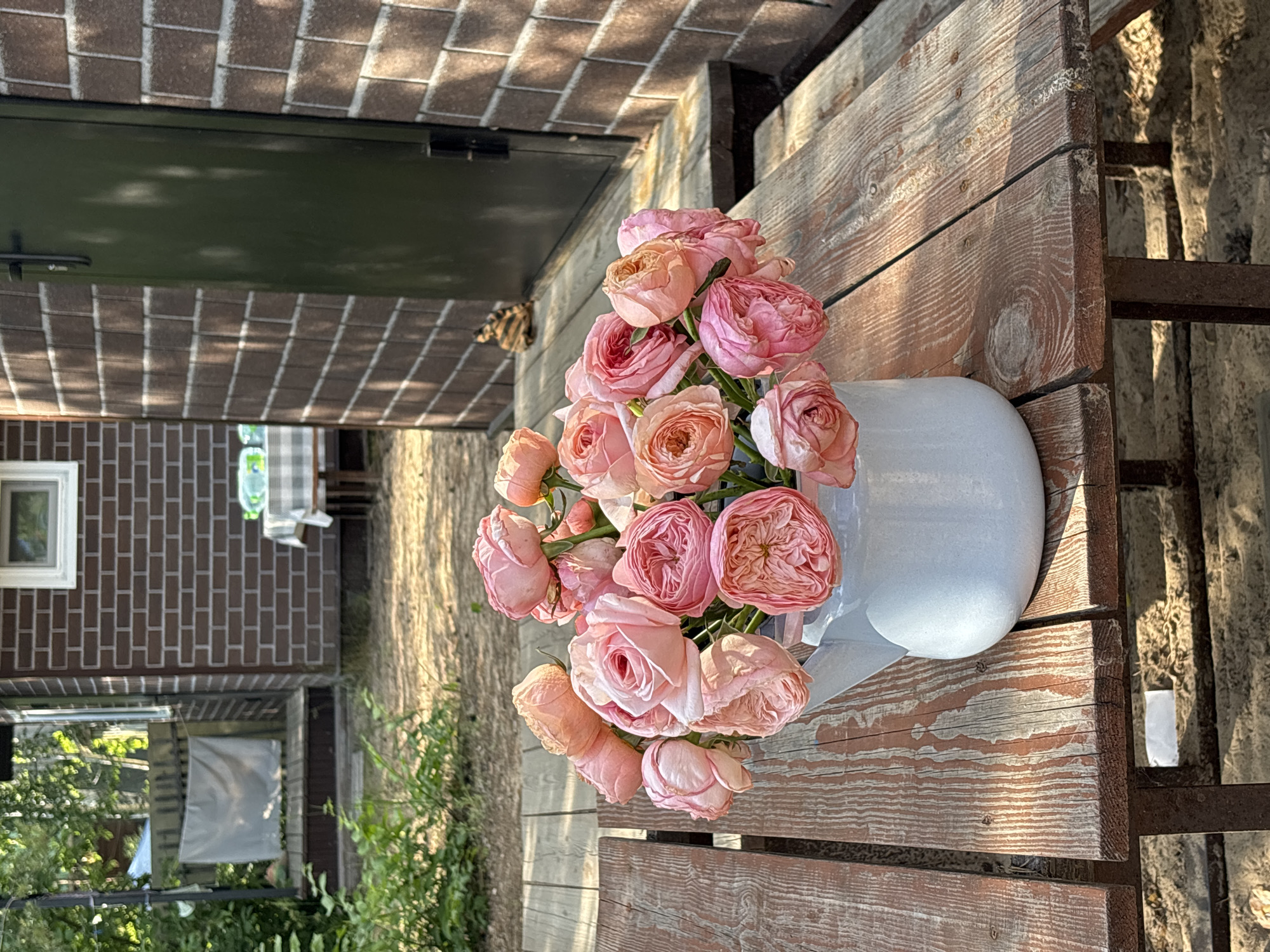 A bouquet of pink roses is arranged in a light blue watering can. The watering can sits on a rustic wooden table. In the background, a brick building with a green door and a window is visible. The scene appears to be outdoors, with greenery and sunlight casting shadows on the table. The location is Kzyl-Bairak, Russia. There are no people in the image.