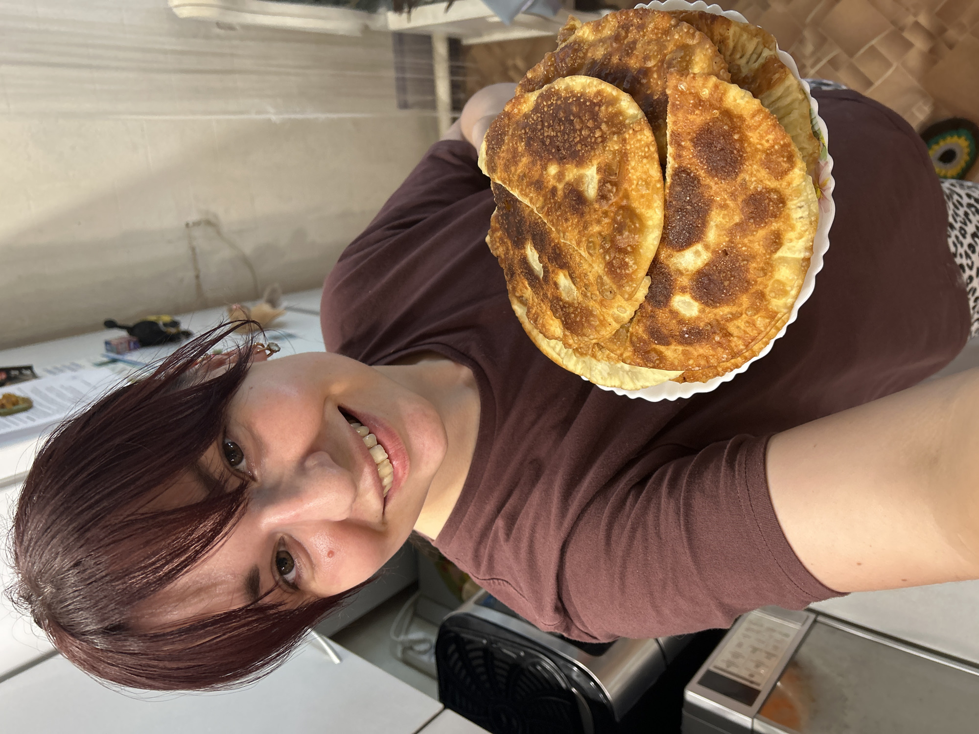 A medium shot shows a young woman with dark red hair and a maroon t-shirt holding a plate of fried pastries. She is smiling and looking at the camera. In the background, a kitchen is visible with appliances such as a refrigerator and an air fryer. The floor is tiled. The overall scene depicts a domestic setting, possibly in the city of Фоки, Russia, where the woman is showcasing freshly made food.