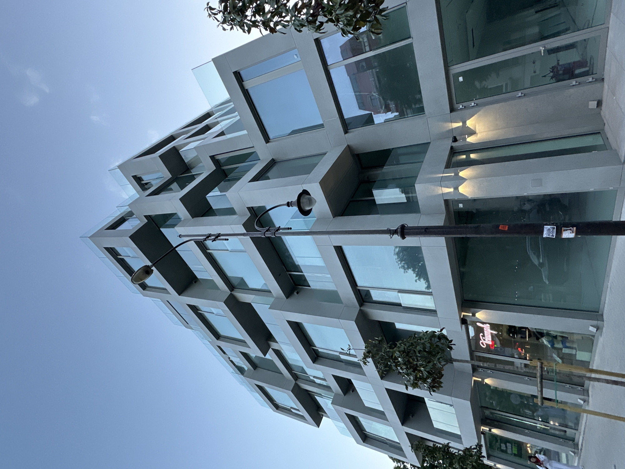 A low-angle shot of a modern, multi-story building with a geometric, box-like facade. The building appears to be a mixed-use structure with both residential and commercial spaces. The windows reflect the sky and surrounding environment. A street lamp is positioned in front of the building. The lower level features a store with a sign that reads "Triumph." Based on the signage and architectural style, the location might be a city center of Bulgaria, possibly Varna. The lighting suggests either early morning or late afternoon. There is also the presence of what appear to be trees. A person is observed walking past the store.