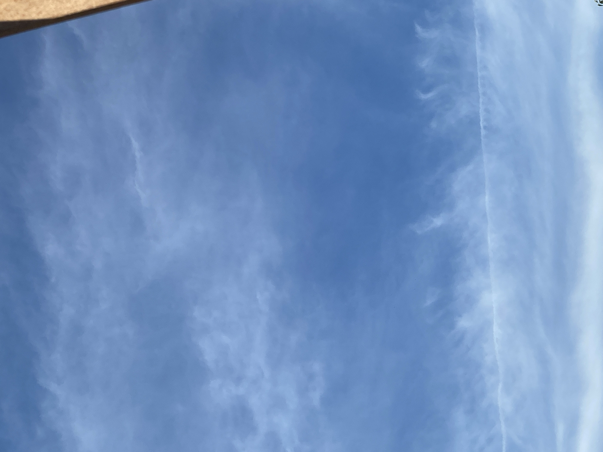 A close-up, vertical, eye-level outdoor shot depicts a vast, clear blue sky with wispy cirrus clouds stretching horizontally across the frame. A faint contrail cuts a thin, white line through the mid-section of the sky. The top right corner of the image shows a sliver of a light brown, vertical object that appears to be a building or structure. The overall impression is a serene, expansive sky.