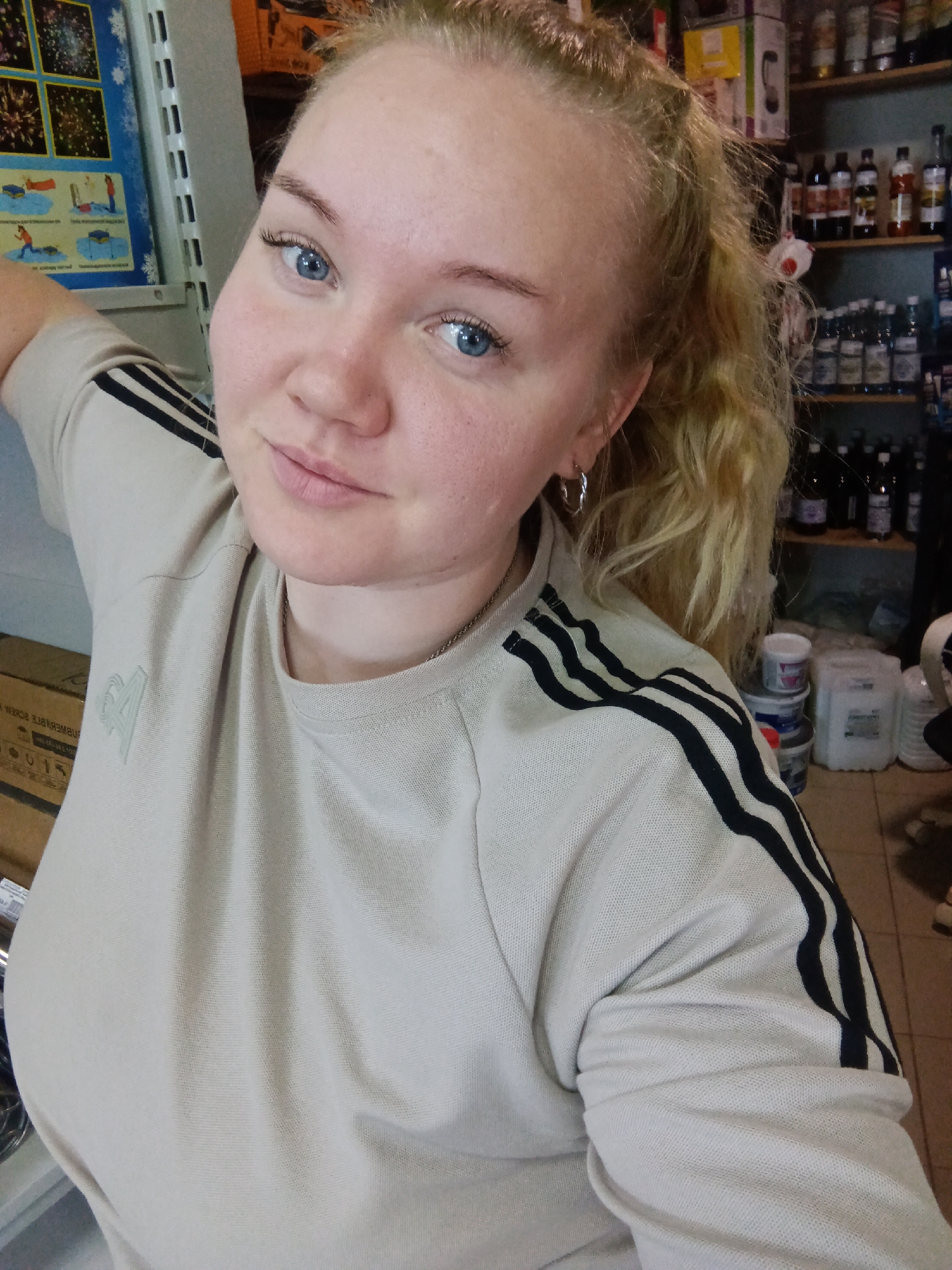 A fair-skinned woman with blue eyes and blonde hair pulled back in a ponytail is posing for a selfie in what appears to be a retail environment, likely a store in Фоки, Russia. She's wearing a light-colored sweatshirt with black stripes on the sleeves. The background is filled with shelves stocked with various items, including bottles, jars, and containers. A sign or poster is visible on a nearby wall. The woman's expression is slightly smiling, with a subtle tilt of her head.
