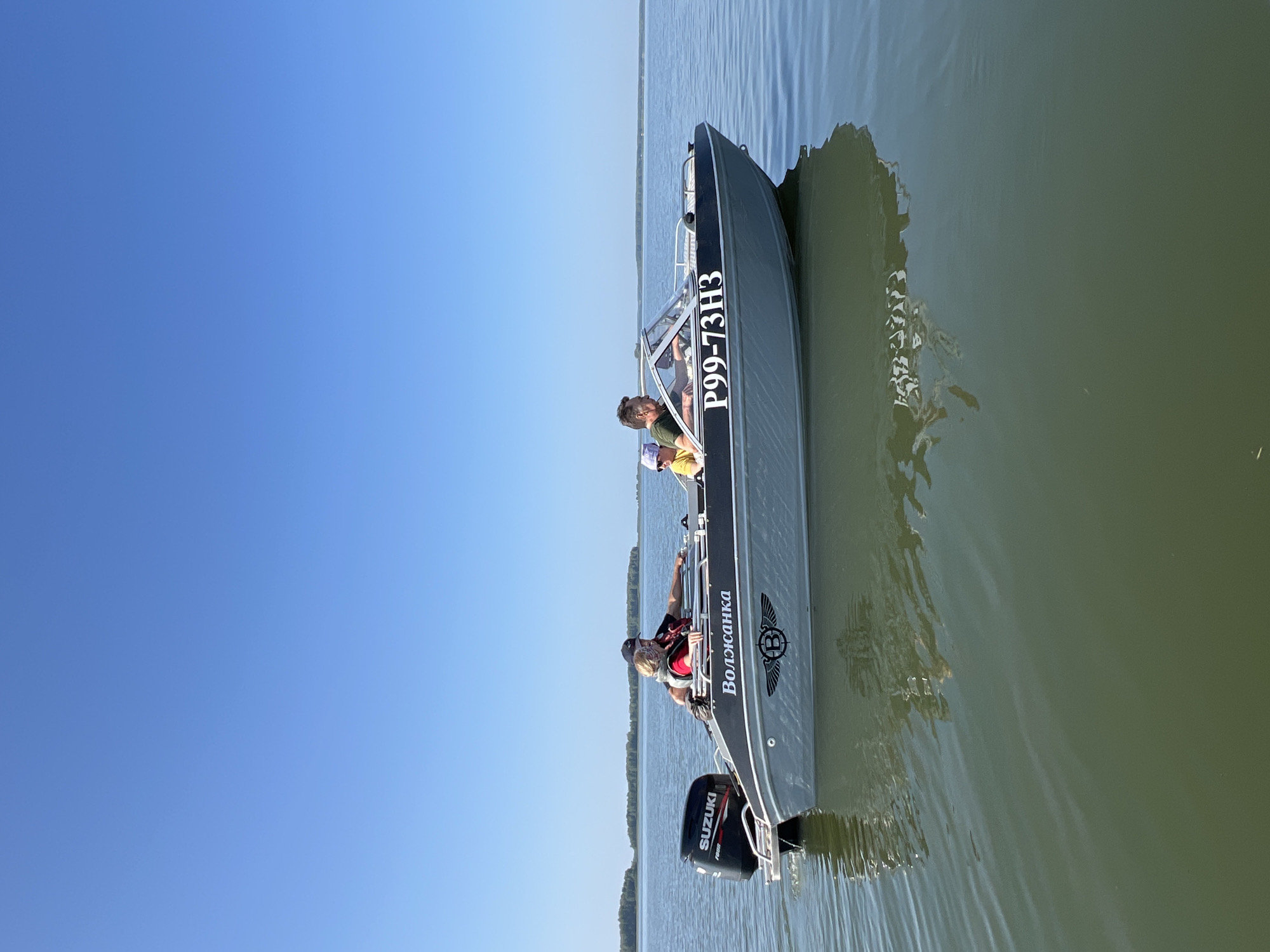 In Novopichugovo, Russia, on a clear, sunny day, a motorboat with three occupants travels across a calm body of water. The boat, gray with black trim, has the name "Волжанка" and the registration number "P99-73H3" painted on its side. A black Suzuki outboard motor is mounted on the stern. Two adults and one child are seated in the boat, all appearing to be enjoying the ride. The adult in the driver's seat is wearing a dark t-shirt and a cap, while the passenger next to them is wearing a red life vest over a light-colored shirt. The child, also wearing a life vest, is seated behind the driver. The background shows a distant treeline under a vast blue sky, reflecting the clear weather conditions.