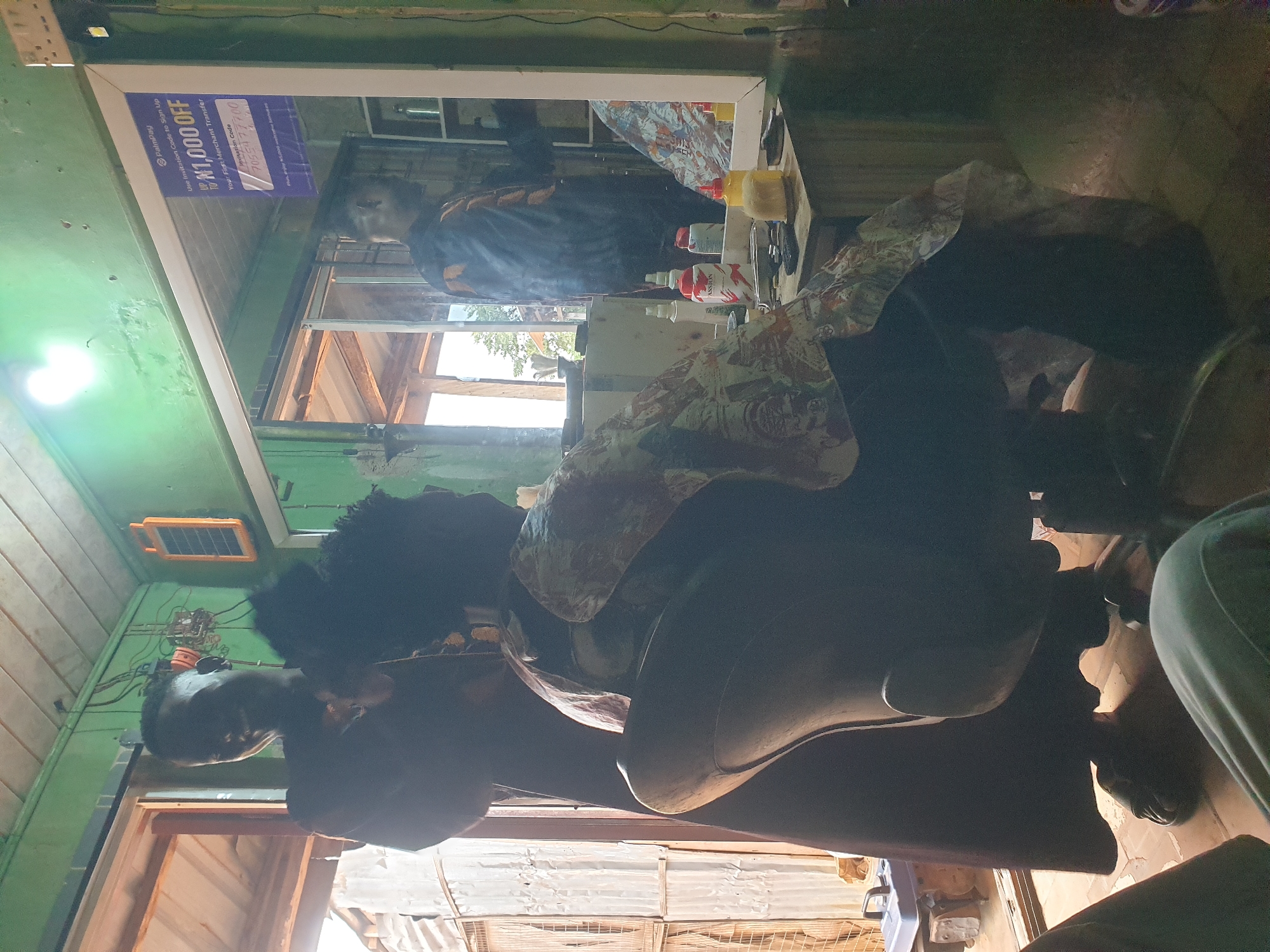 In Oluawo, Nigeria, a dimly lit barber shop interior is depicted, with a focus on a seated individual undergoing a haircut. A barber, whose face is obscured by shadow, stands behind the seated person, holding a tool near their head. The seated individual is draped in a patterned cloth. In the background, a mirror reflects a man in a dark patterned smock standing near a shelf with bottles, and a poster advertising a discount for a digital payment service. The shop has a rough, corrugated metal exterior wall visible through an open doorway and a green-painted interior with exposed wiring and a small solar panel. A generator is partially visible near the doorway. The floor appears to be tiled.