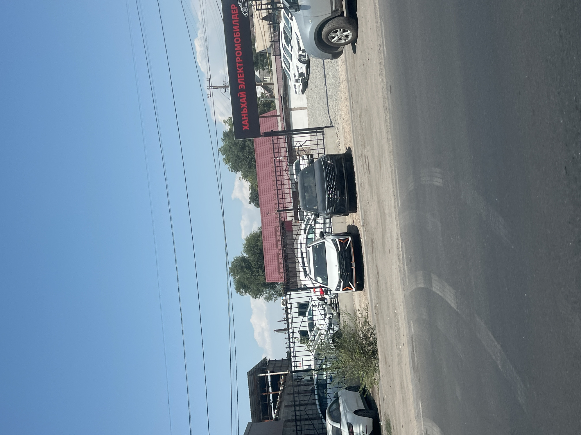A sunny, clear sky is visible above a car dealership in Bishkek, Kyrgyzstan. The dealership has a dark sign with red text that reads "ХАНЬХАЙ ЭЛЕКТРОМОБИЛДЕР" (Hanxai Electromobiles) and an image of a car. Several white electric cars are parked in a row behind a black metal fence. In the foreground, a black SUV and a white sedan are parked on a dirt lot. A grey SUV is visible on the right side of the image. A road with tire marks is in the foreground, and trees and buildings are visible in the background.