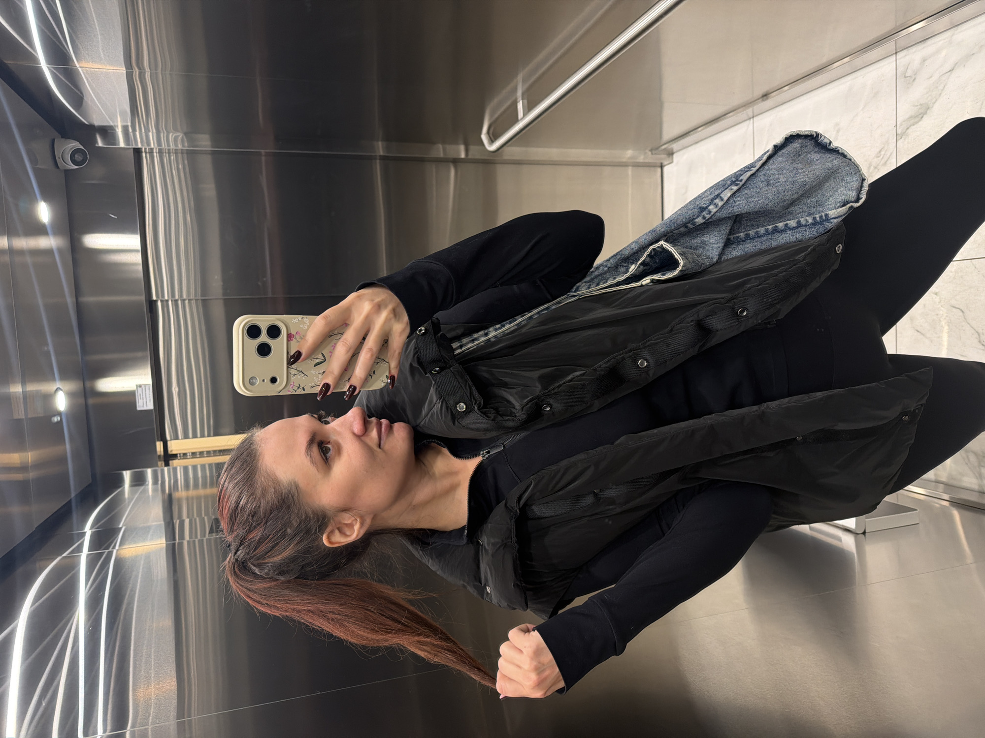 The image depicts a female individual taking a selfie within a modern elevator in Belgrade, Serbia. The elevator interior features polished, reflective stainless steel panels on the walls and ceiling, which show horizontal reflections of overhead lighting. A white dome-shaped security camera is mounted in the upper left corner of the scene. The floor is covered with light-toned tiles displaying subtle grey veining. A silver-colored horizontal handrail is visible on the right wall.
The subject is a female with reddish-brown hair styled in a high ponytail. She is dressed in an all-black ensemble consisting of a long-sleeved quarter-zip top, a padded black vest, and black leggings. Her left hand holds a light-colored smartphone with a floral-patterned case, used to capture the image. Her nails are manicured with dark polish. Her right hand is positioned near her head, gently gripping a section of her ponytail. A light-wash denim bag is visible, slung over her left shoulder and hanging down her left side. Her gaze is directed towards her right, away from the phone's camera lens.