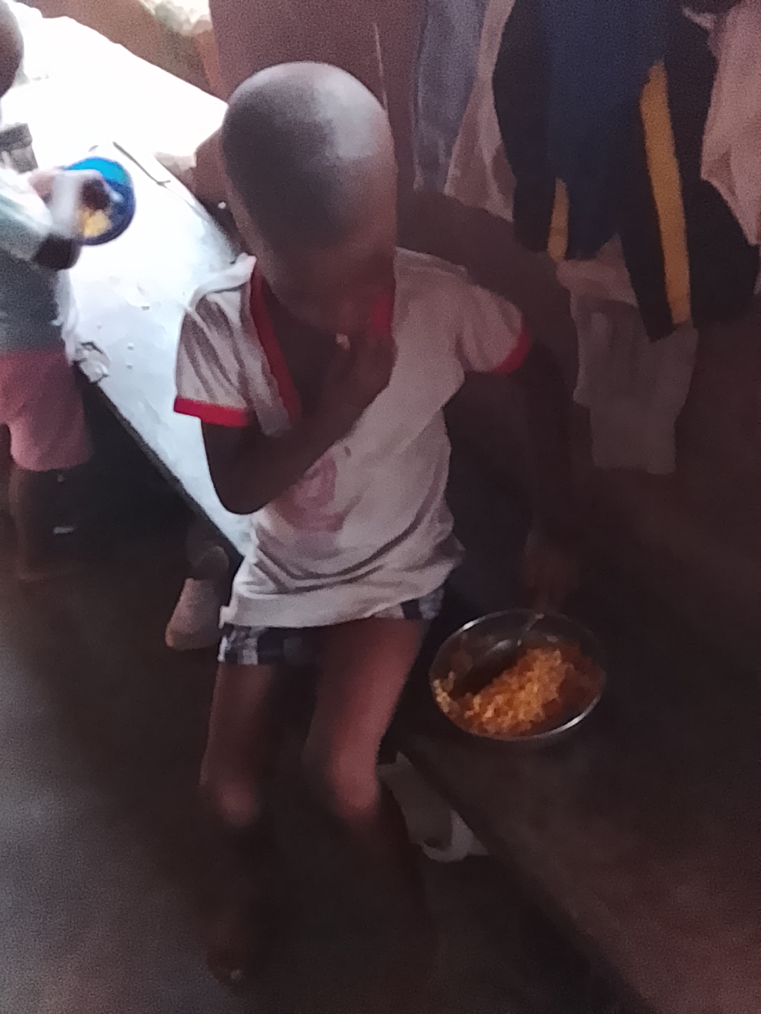 The image captures a scene indoors in Nkpor, Nigeria, featuring multiple individuals, primarily children, during what appears to be a mealtime. The central subject is a child with a shaved head, wearing a white short-sleeved t-shirt with red trim and plaid shorts. This child is seated or kneeling next to a wooden surface, where a metal bowl filled with an orange-brown, grain-like food (possibly rice or a similar dish) and a spoon are placed. The child's left hand is near their mouth, suggesting they are eating or about to eat. An adult's arm is visible, seemingly resting on or around the central child's back, indicating a supportive presence.
To the left, another child is partially visible, wearing pink shorts and holding a blue cup or small bowl, standing near a light-colored surface, possibly a table or bench. Further details of other individuals are obscured, though parts of legs and clothing are discernible in the background, including a black garment with a yellow stripe. The lighting in the room is dim, casting a somewhat muted and intimate atmosphere. No text is visible in the image. The scene conveys a simple, everyday moment of sustenance within what appears to be a domestic or communal setting.