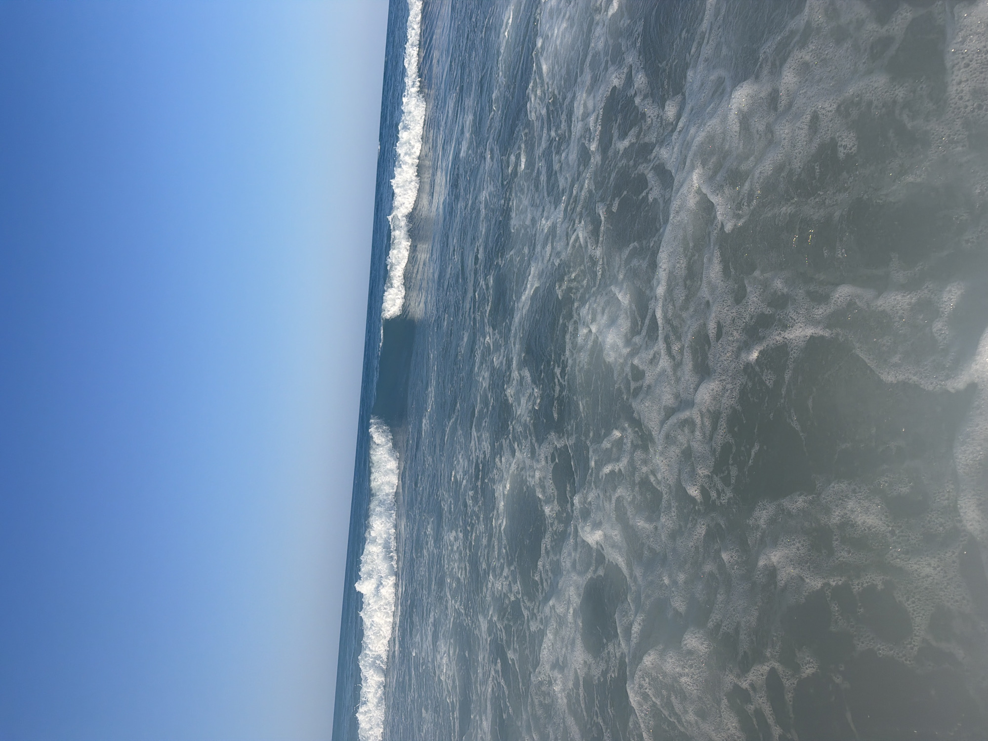 The image depicts a serene seascape under a clear blue sky. The foreground is dominated by foamy, agitated seawater, indicative of waves that have recently broken or are in the process of receding. These white froth patterns create a dynamic texture against the darker blue of the deeper water. Further out, several waves are visible, some cresting with white foam, stretching across the frame towards the horizon. The horizon line is distinct, separating the deep blue of the sea from the pale blue of the sky. The scene is devoid of any human presence, boats, or man-made structures, suggesting a natural and undisturbed environment. This image could be captured at any coastal location. However, if specifically to be located in Georgioupoli, Greece, it would be a view of the Aegean Sea from its coastline.