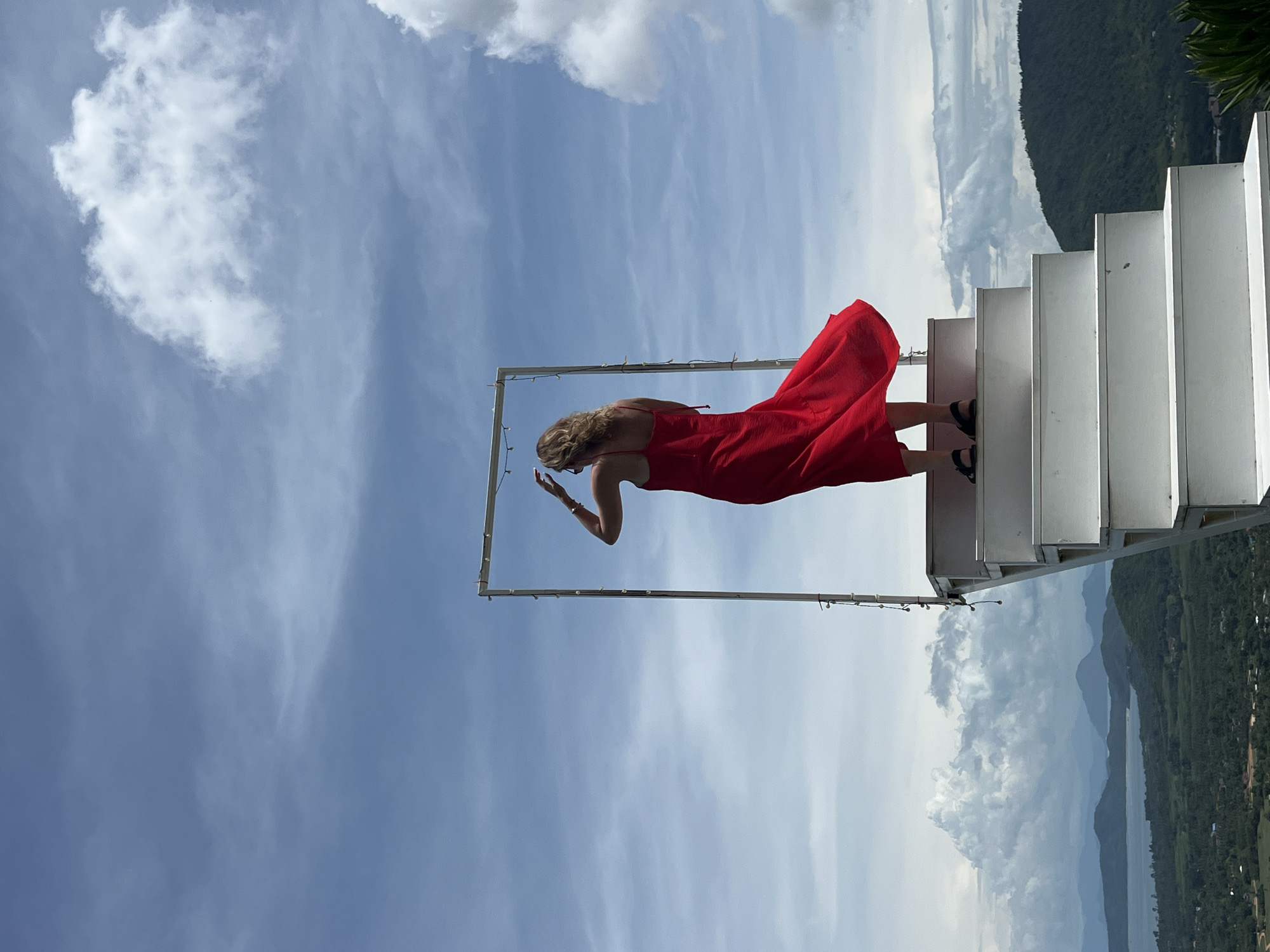 A woman stands on a white staircase, facing away from the camera. She is wearing a red dress with spaghetti straps and her hair is blonde. The dress is flowing in the wind. She has her hands on her head, adjusting her hair. She is wearing black sandals. In front of her, there is a rectangular frame, possibly an art installation or a prop. The staircase leads up to this frame. The background is a vast sky with white clouds against a blue sky. Below the staircase, a landscape unfolds, featuring lush green hills, a body of water that appears to be a lake or bay, and mountains in the distance. The location is identified as Baan Thurian, Thailand. The overall scene suggests an outdoor scenic viewpoint or a tourist attraction.