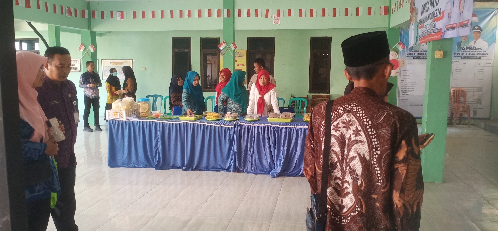 In Tamansari, Indonesia, a group of people are gathered in what appears to be a community hall decorated with Indonesian flags. In the foreground, a person wearing a batik shirt faces away from the camera, holding a phone. To the left, a couple stands near an entrance. The central area features a table covered with a blue tablecloth, laden with various food items displayed in trays and jars. Several women, some wearing hijabs, are positioned behind the table, interacting with the food and potentially serving visitors. In the background, posters with text are affixed to the light green walls, and chairs are visible. The flooring is tiled. The scene suggests a local event or exhibition in Tamansari.