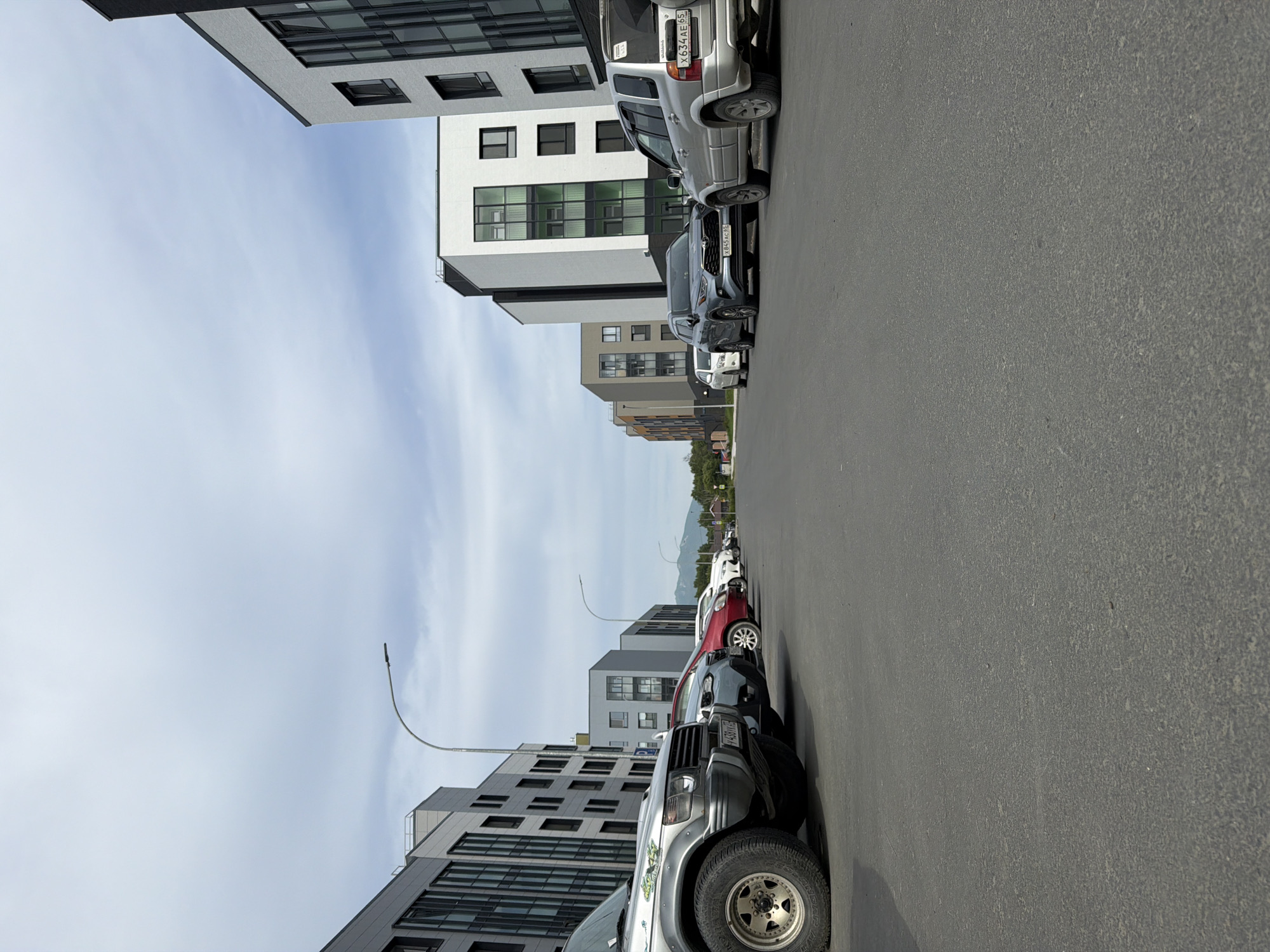 A low-angle shot captures a street scene in Yuzhno-Sakhalinsk, Russia, featuring modern apartment buildings and parked cars. The foreground is dominated by asphalt pavement, with the left side of the image showing a portion of a silver SUV. Several other cars are parked in a row on the left, including a red sedan and a blue SUV. On the right, a silver SUV with a license plate that reads "X634AE 65" is prominently displayed. Further down the street, more cars and buildings are visible. The buildings are contemporary in design, with a mix of white, gray, and dark facades, featuring numerous windows. A street lamp arches over the road on the left. In the distance, a hazy mountain range is discernible against a cloudy sky. The scene appears to be a residential or commercial area, likely a parking lot or street adjacent to apartment complexes. No people are visible in the image.