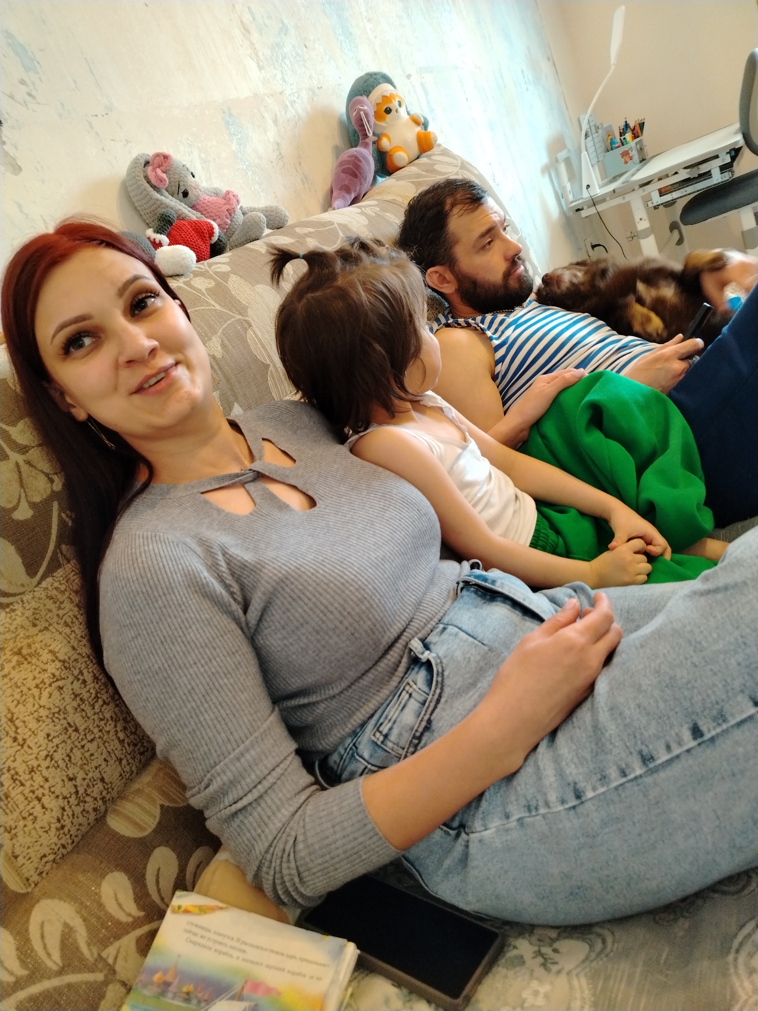 A woman, a child, and a man are on a patterned couch. The woman is wearing a gray long-sleeved shirt and jeans. The child is wearing a white tank top and green pants. The man is wearing a striped blue and white tank top. A dog is lying on the man. Stuffed animals are on the couch behind them. A desk and chair are in the background. A book and a black phone are on the couch next to the woman. The setting is indoors, presumably a home. There is no evidence in the image to indicate the city of Noviy, Russia.