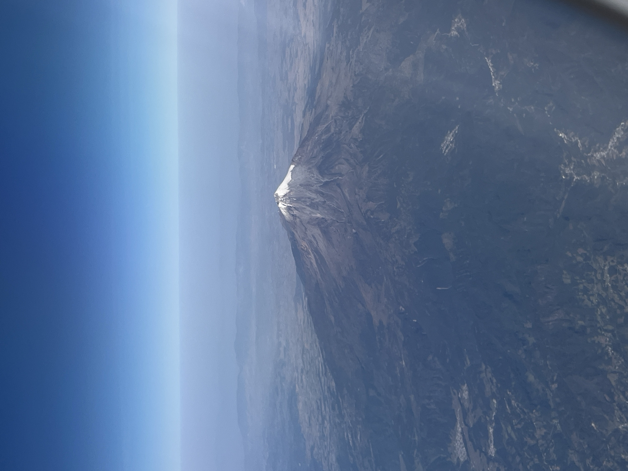 An aerial photograph captures a majestic snow-capped volcano rising from an expansive, rugged landscape under a clear daytime sky. The image, likely taken from an aircraft, looks down on the terrain, with the horizon line visible in the upper portion of the frame.
The central subject is a conical, dark-colored volcano, its summit clearly defined by a brilliant cap of white snow or ice. The mountain's slopes are deeply furrowed and textured, appearing rocky and barren in places, with variations in shade indicating valleys and ridges. Below the snow line, the mountain transitions into a vast, undulating terrain, characterized by muted tones of green, brown, and grey, suggesting a mix of sparse vegetation and exposed earth. Atmospheric haze softens the distant landscape, creating layers of receding hills and plains.
The sky occupies the upper left portion of the image, displaying a gradient of blue, darkening towards the upper edge and lightening to a pale, almost white hue near the hazy horizon. The overall impression is one of immense scale and natural grandeur. Given the context of Tenancingo, Mexico, the prominent snow-capped peak is very likely Nevado de Toluca (Xinantécatl), a well-known stratovolcano in the region. There are no people or discernible text in the photograph. The clear skies and bright illumination suggest it is mid-day.