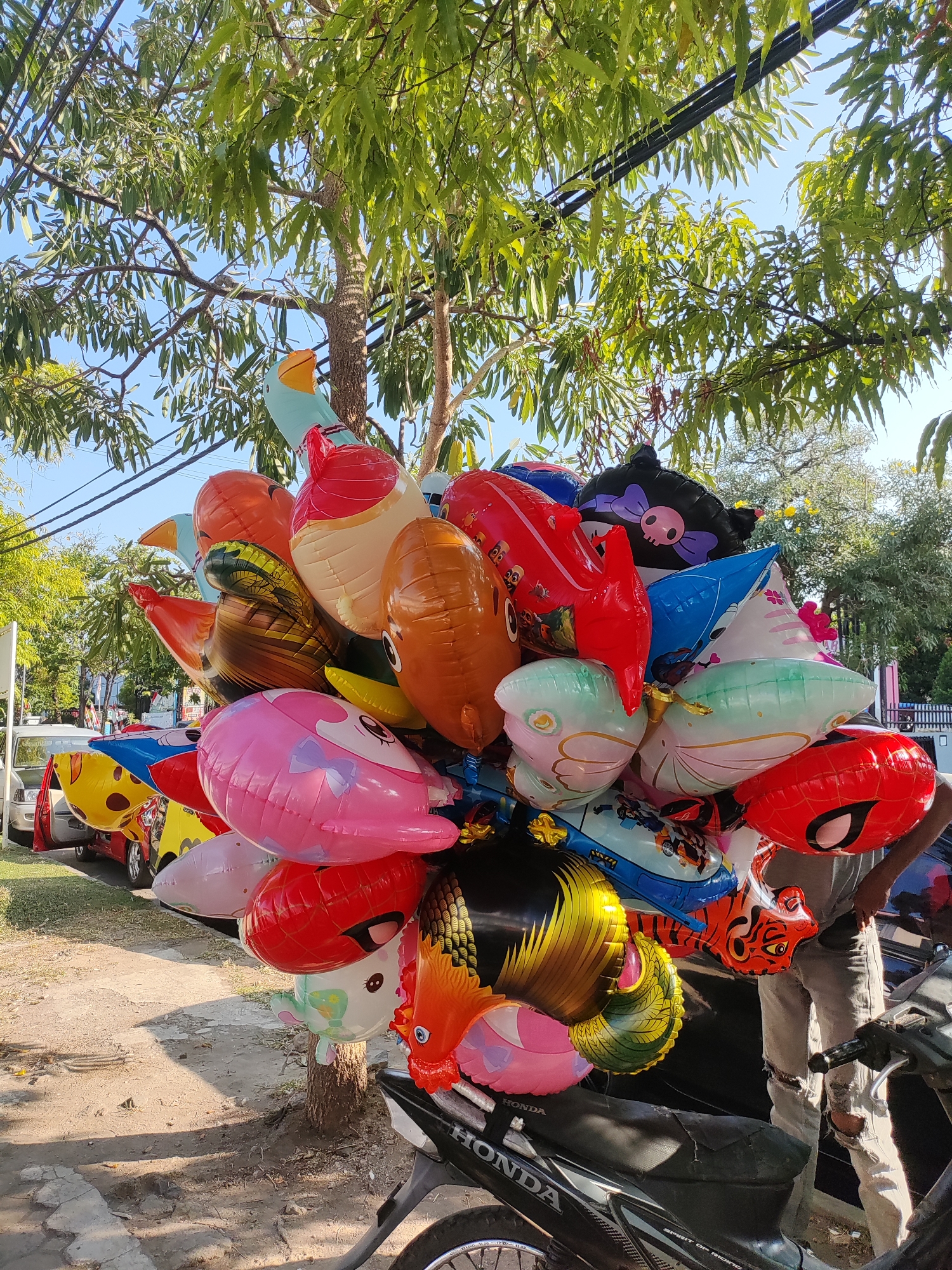 A close-up, eye-level, medium shot captures a large bunch of colorful, inflated balloons tied to a tree trunk. The balloons, in various animal and character shapes, are predominantly red, orange, blue, pink, and black, with some featuring printed designs of cartoon faces, tigers, and superheroes. They are arranged in a dense cluster, obscuring the trunk and branches of the tree. Overhead, lush green leaves of the tree create a canopy, with glimpses of a clear blue sky and black power lines visible through the foliage.
In the lower right portion of the frame, a person is partially visible, wearing ripped light-colored jeans and a dark garment. Their hands are engaged with the balloons, suggesting they are holding or adjusting the bunch. The balloons are anchored to the handle of a black motorcycle, the brand "HONDA" is visible on its side. The motorcycle is parked on a dirt and gravel surface, with a portion of a paved sidewalk to the left.
In the background, a white car with its door open is parked on the grassy area beside the road, and other vehicles are visible further down the street. A black metal fence is also present in the distance. The overall setting appears to be a roadside or street vendor location in a tropical or sub-tropical environment, likely in the city of Cirebon, Indonesia, given the context of the image. The lighting indicates it is daytime, with sunlight casting shadows on the ground.