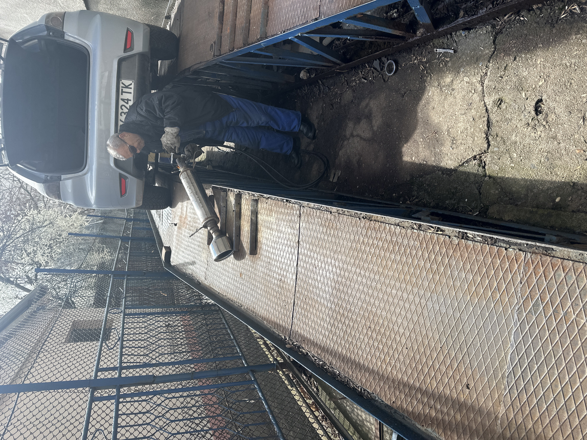 The scene depicts an outdoor car repair area in Stara Zagora, Bulgaria. A single adult male, wearing dark sunglasses, a dark jacket, blue work trousers, and light-colored work gloves, stands in a concrete service pit. He is positioned beneath the rear of a silver hatchback car (Bulgarian license plate CT 6324 TK), which is elevated on rusty metal diamond-plate ramps. The man is actively engaged in work on a metallic pipe, likely an exhaust component, using a tool that is emitting sparks, suggesting cutting or welding. An additional, similar metallic pipe with a chrome tip rests on the left ramp. The ground in the service pit is concrete, cracked, and littered with debris and wires. To the left, a blue metal chain-link fence runs parallel to the ramps, behind which a light-colored building with a small window is visible. Bare tree branches, some with white blossoms, are seen in the background. The area is illuminated by bright sunlight, casting strong shadows.