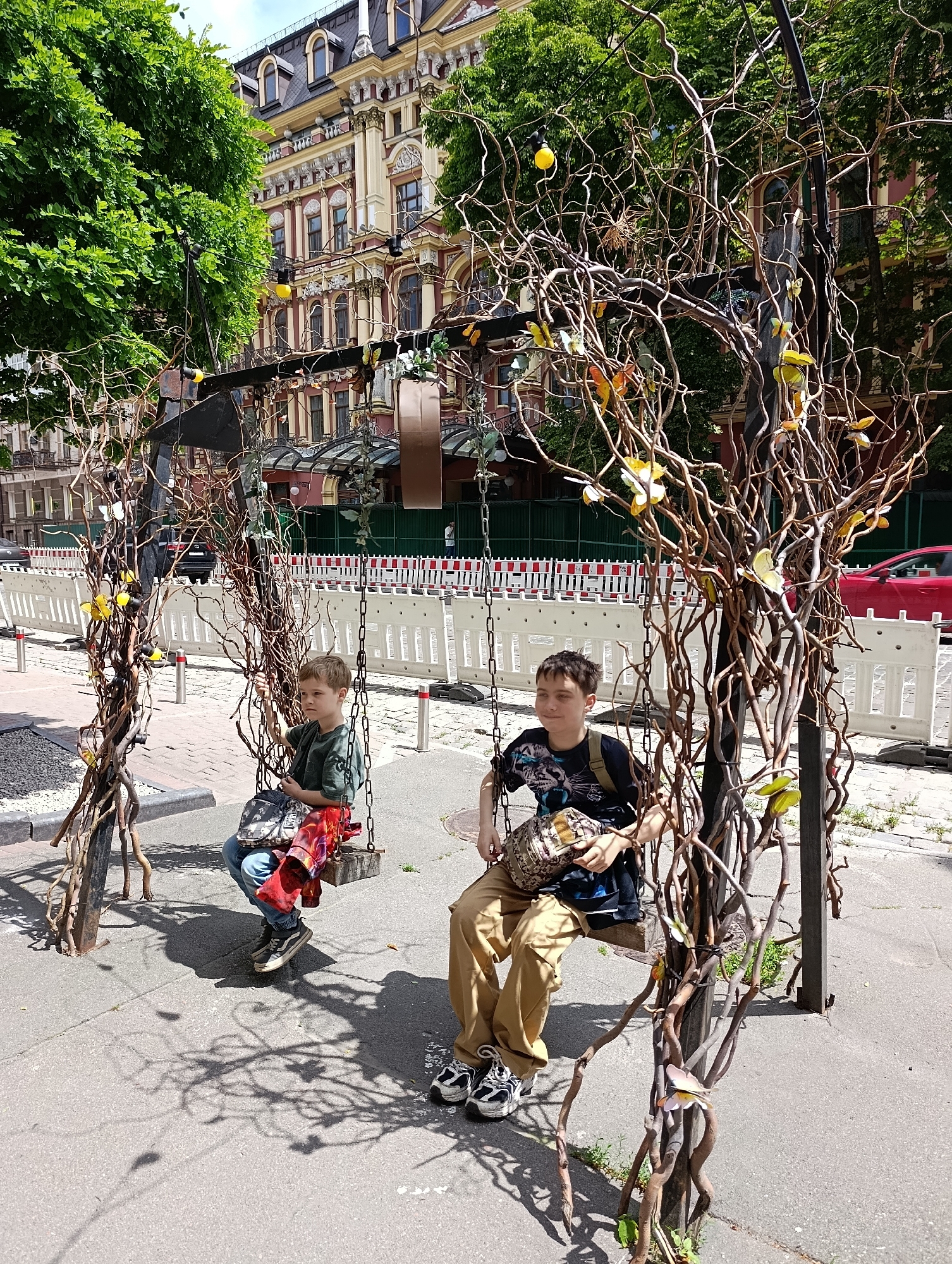 Two young boys are sitting on swings, which are part of a decorative tree-like structure adorned with artificial butterflies and lights. The swings themselves appear to be made of wooden planks suspended by chains. The boy on the left is wearing a green t-shirt and jeans, while the boy on the right is wearing a black t-shirt with a tiger print and khaki pants. Both are holding bags on their laps.
The scene is set outdoors on a paved area, possibly a sidewalk or a pedestrian zone. Behind the swings, there's a white barrier fence, suggesting ongoing construction or event setup. Beyond the fence, a red car is visible, and further back, an ornate, multi-story building with classical architectural elements stands. Lush green trees frame the scene on the left. The ground shows intricate shadows cast by the swings and the decorative tree structure. The overall impression is a sunny day in an urban environment, with elements of public art and possibly a public space that is being prepared or has been adapted for an event. Based on the architectural style of the building and the presence of similar decorative elements observed in other public spaces, the location is likely Kyiv, Ukraine, possibly in the Podil district, which is known for its historical buildings and pedestrian-friendly streets.