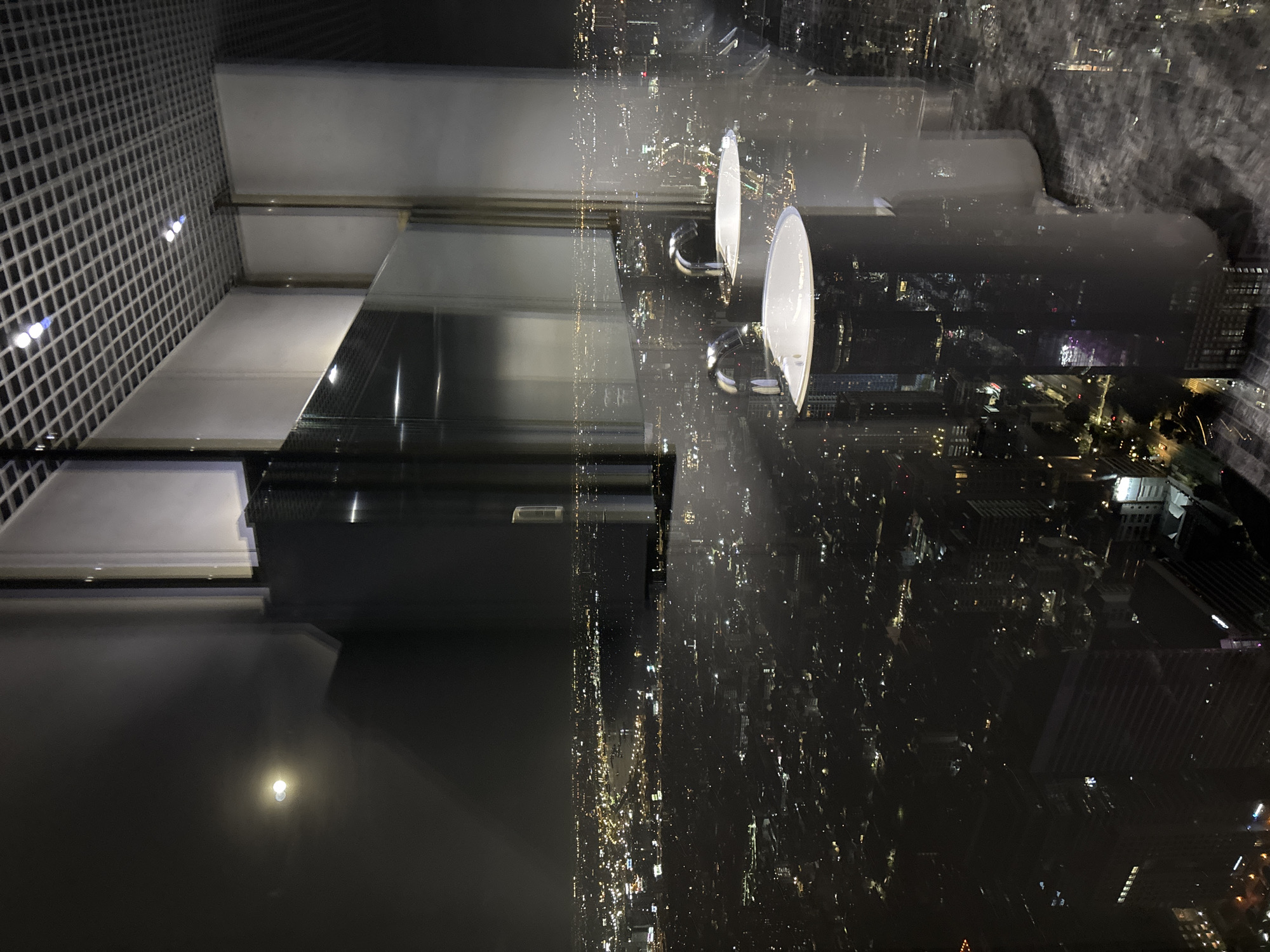 The image, rotated counter-clockwise, depicts a night view from a high-rise building in Bangkok, Thailand, looking out over the illuminated cityscape. The scene combines elements of an indoor space with reflections on a large window and the expansive urban panorama outside.
In the foreground, to the left, is a white or light-colored gridded structure, possibly a decorative wall or a ceiling panel, with several small, bright blue-tinged lights embedded within its squares. The central and right foreground is dominated by a large, dark, highly reflective surface, likely a table or counter. On this surface, or positioned near the window, are two modern, elegant lamps with white, circular tops and shiny, curved chrome bases. These lamps and other indoor details are clearly reflected on the expansive window, overlapping with the view of the city.
Beyond the reflections and through the window, a vast metropolis stretches into the distance, illuminated by countless electric lights from buildings, streets, and vehicles. The night sky is dark, contrasting sharply with the bright points of light across the urban landscape. Silhouettes of distant buildings are faintly visible amidst the glow. The overall impression is one of observing a vibrant city at night from a high vantage point, such as a sky bar, restaurant, or observation deck. No people are visible, and there is no discernable text.