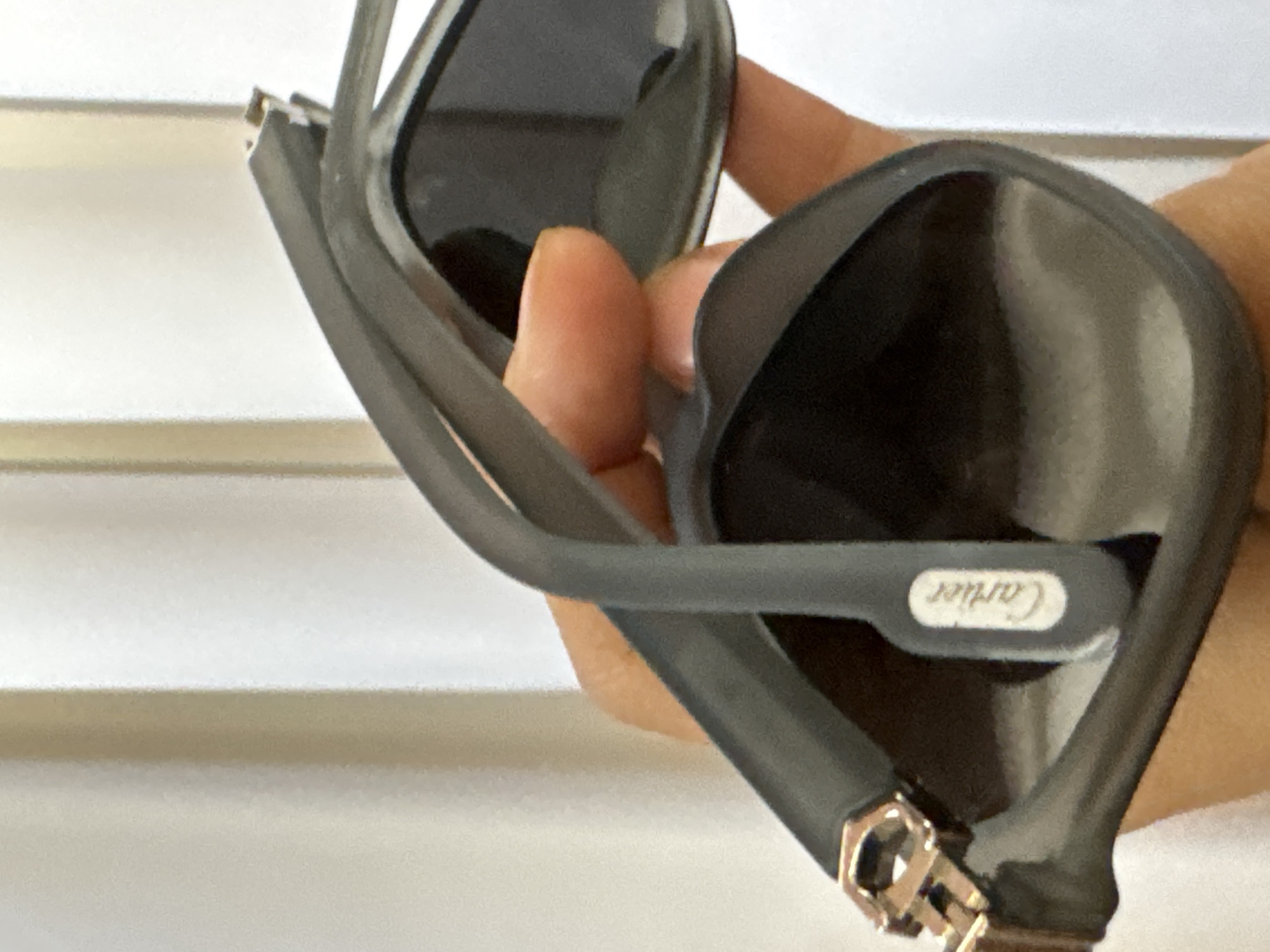 The image shows a close-up of a pair of dark gray Cartier sunglasses being held by a hand. The right lens is angled towards the viewer, displaying its dark tint. The temple of the glasses features the Cartier logo in white. The hinge of the right temple is visible, showing a gold-colored metallic detail. The background is a blurred white, possibly a wall or surface. No information suggests the image was taken in Davao City, Philippines.