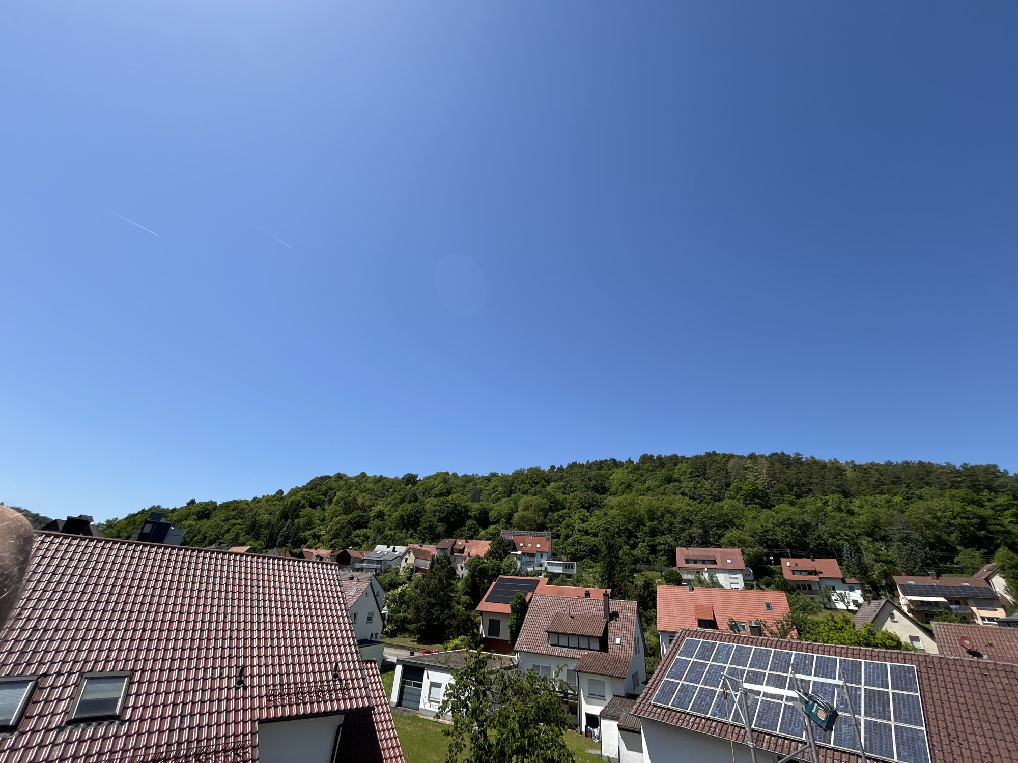The image captures a daytime view of a residential area in Mönsheim, Germany. The perspective is elevated, likely from a building or high point. In the foreground, rooftops covered in red clay tiles are prominent, some fitted with solar panels. One rooftop has a metal ladder resting against it. The midground features a dense mix of residential buildings interspersed with greenery. Beyond the houses is a heavily wooded hillside, forming a natural backdrop to the urban landscape. The sky is clear and bright blue with a contrail visible.