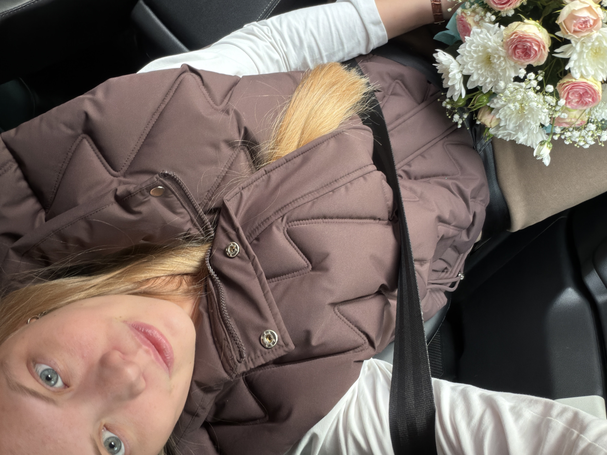 A young woman is captured in a close-up, low-angle shot, seemingly taken from inside a car in Uspenskii, Russia. She has blonde hair and light blue eyes, looking directly at the camera with a soft, pleasant expression. She is wearing a white long-sleeved top underneath a dark brown, quilted puffer vest. A black seatbelt crosses diagonally across her chest and shoulder. Part of her blonde hair spills over the top of the vest.
Next to her, on what appears to be a car seat or armrest, rests a bouquet of fresh flowers. The bouquet includes light pink roses, white chrysanthemums, and small white baby's breath, wrapped in beige paper with a hint of light blue ribbon or paper visible. A watch with a brown leather strap is visible on her left wrist, near the flowers. The car's interior features dark upholstery, visible as black seats around her. The image conveys a casual, personal moment, possibly on a cool day given the puffer vest. No text is visible in the image.