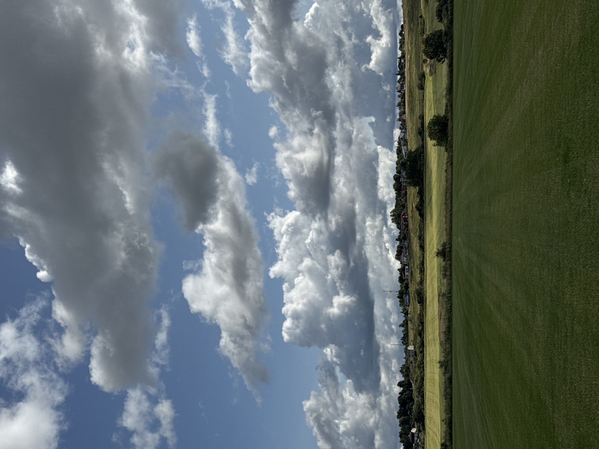 This is a landscape photograph taken in Kempelevo, Russia, under a dramatic sky filled with cumulus clouds. The foreground is dominated by a vast, neatly mown green field, its surface showing subtle striations from the cutting. Beyond this field, a gentle slope rises, featuring a strip of lighter green, possibly a different crop or pasture, bordered by a thin line of trees and bushes. Further up the slope, a small village or settlement is visible, with various houses and buildings scattered across the ridge, interspersed with trees. The buildings are predominantly red and white. The sky above is a vibrant blue, broken by large, fluffy white and grey clouds, some casting shadows on the landscape below. The overall impression is of a rural, open landscape on a partly cloudy day. There are no people or animals visible in the image.