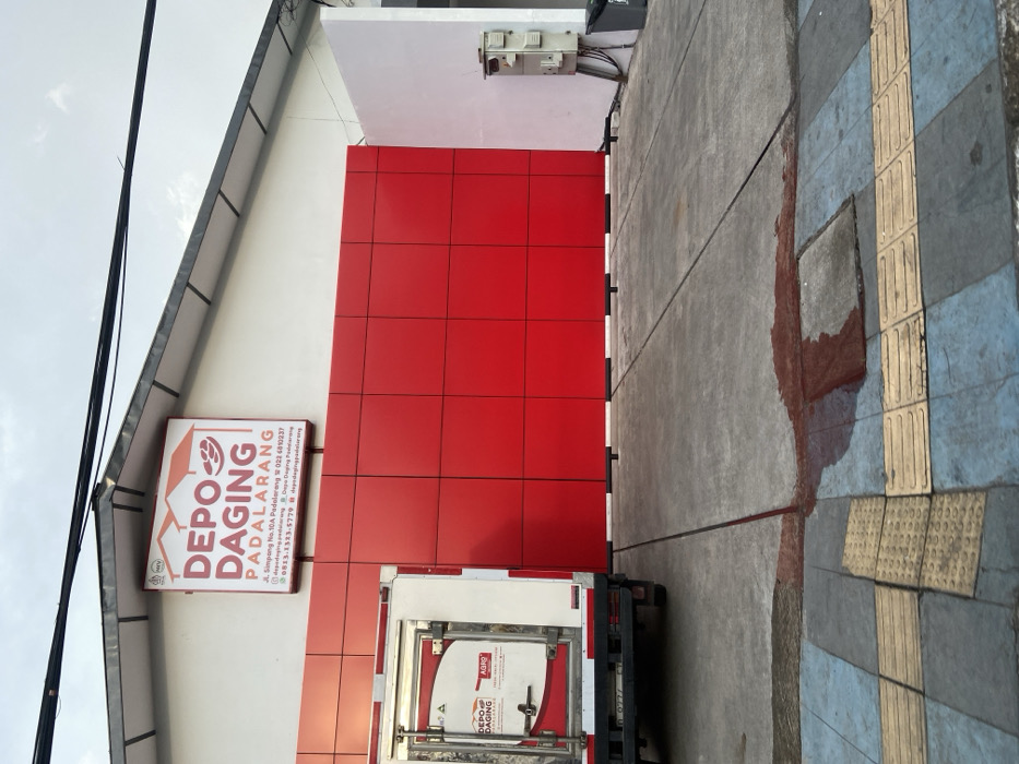 A white delivery truck with a red "DEPO DAGING" logo is parked in front of a red-painted building. The building has a sign that reads "DEPO DAGING PADALARANG" in large white letters. The sign includes the address and phone number. The truck is facing the building, which is located in Padalarang, Indonesia, at coordinates -6.85 latitude, 107.497 longitude. The truck is parked on a concrete pad in front of the building. There is a small patch of red liquid on the ground near the building. The environment is a commercial area with a parking lot and a sidewalk. There are no people present in the image.