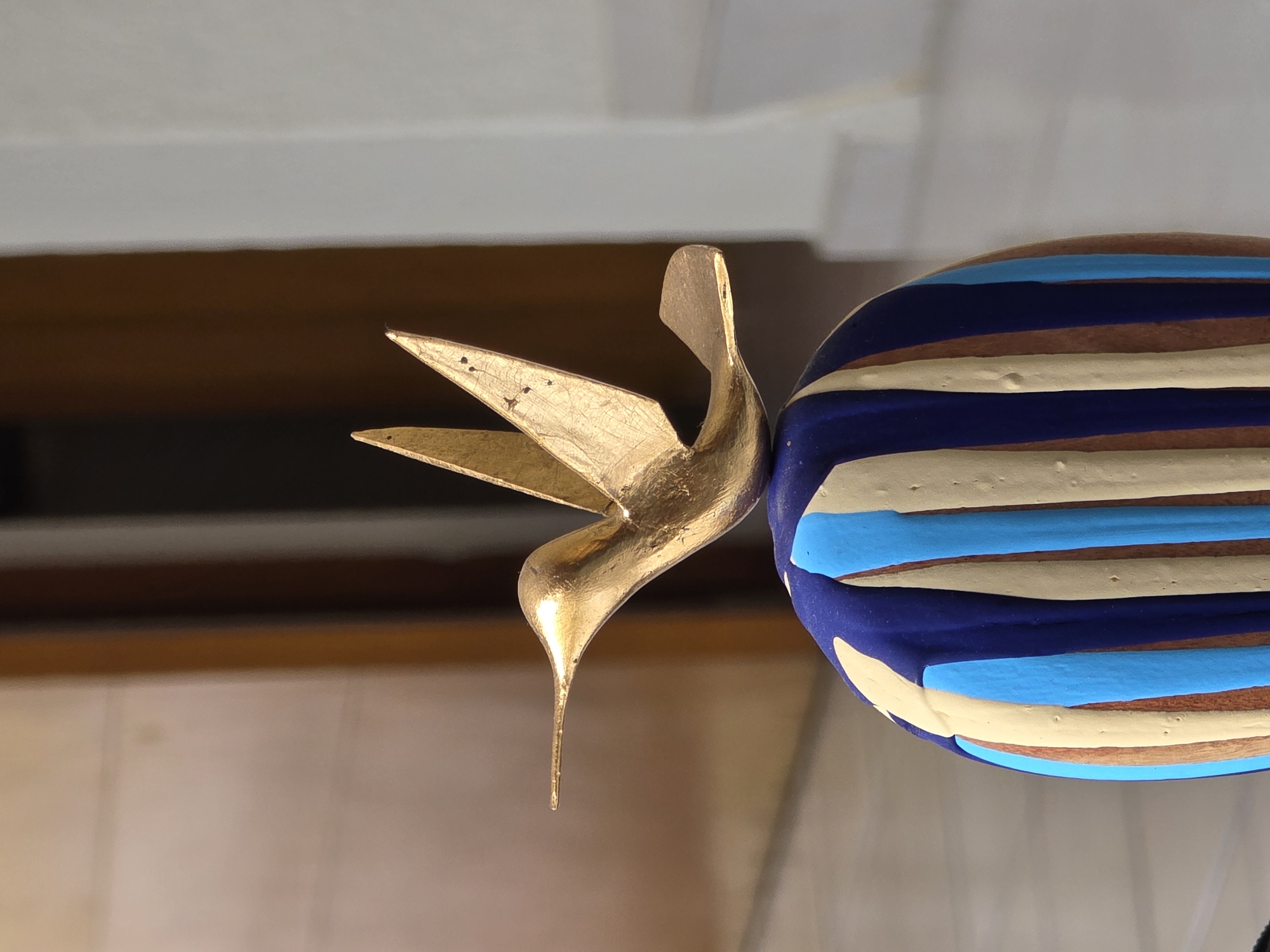 The image displays a close-up of two decorative objects, set against a blurred, indistinct background.
The primary subject on the left is a stylized golden hummingbird figurine. It appears to be made of metal or a material coated with gold leaf or metallic paint, exhibiting a slightly textured, somewhat distressed surface with minor irregularities and a few dark specks. The hummingbird is captured in mid-flight pose, with its wings spread upwards and its long, slender beak pointing downwards. Its form is sleek and minimalist, emphasizing movement.
To the right, partially visible, is a spherical object adorned with horizontal stripes. These stripes are hand-painted and somewhat uneven, featuring alternating bands of dark blue, light blue, cream or light yellow, and a reddish-brown or terracotta hue which might be the base material of the object. The texture of the painted stripes appears slightly raised, indicating a thick application of paint or glaze.
The background is softly blurred, showing a horizontal division. The upper portion is a muted light gray or off-white, while the lower section transitions from a medium brown to a lighter tan, possibly indicating a wall and furniture or another architectural element in an indoor setting. The lighting is diffuse, casting subtle shadows and highlighting the metallic sheen of the hummingbird.
No discernible activity or event is taking place; the image is a still life of decorative items. There is no visible text. Given the context of Puebla, Mexico, the hand-painted striped sphere could be an example of local artisan pottery or Talavera-style ceramics, though the specific pattern isn't strictly traditional Talavera. The golden hummingbird figurine complements the decorative theme.