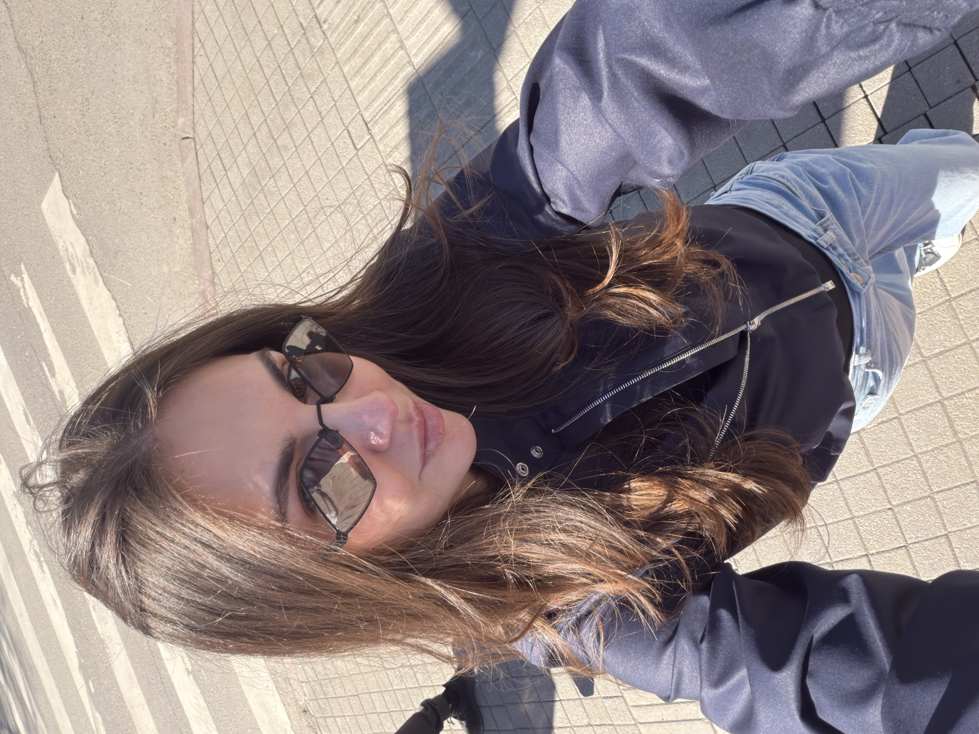 An overhead, rotated selfie depicts a young woman outdoors in Krasnoyarsk, Russia, on a bright, sunny day. The image is oriented with the woman's head at the bottom left and her body extending towards the upper right. She has long, dark brown hair with lighter brown highlights, styled loosely around her shoulders. She is wearing black sunglasses with mirrored lenses, which obscure her eyes. Her lips are slightly parted.
She is dressed in a dark navy or black zip-up jacket over a dark top, paired with light blue denim jeans. A small part of white sneakers is visible near her feet at the bottom right of the frame. Her right arm, covered by the jacket, is raised, suggesting she is holding the camera.
The background primarily consists of various types of paved surfaces. To the left, there's a light-colored, patterned sidewalk made of square or rectangular tiles. To the right, another section of pavement features smaller, darker square blocks. Parts of the ground show a darker asphalt-like surface with a white painted line, possibly indicating a road or crosswalk. Strong sunlight casts distinct, long shadows across the ground and the woman's clothing, indicating either morning or late afternoon. There is no visible text in the image.