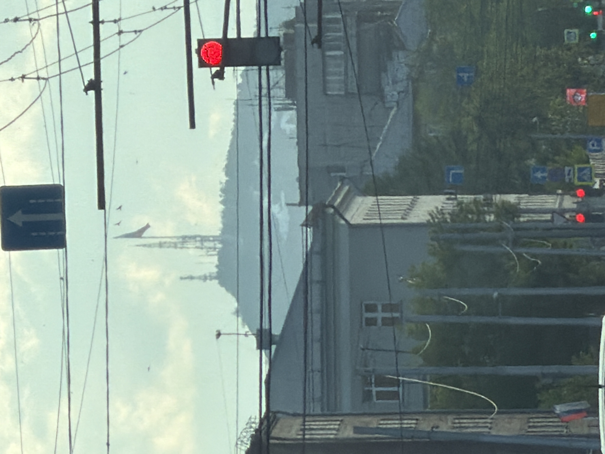 The image shows a street scene in Krasnoyarsk, Russia. A red traffic light is visible in the foreground. A blue directional road sign pointing left is also present. Utility poles and wires crisscross the sky. In the distance, a large body of water is visible, with a land mass beyond. An airplane is flying in the sky. Several buildings, including an older, light-colored building with multiple windows, line the street. Other traffic lights can be seen in the distance. Trees and other vegetation are present.